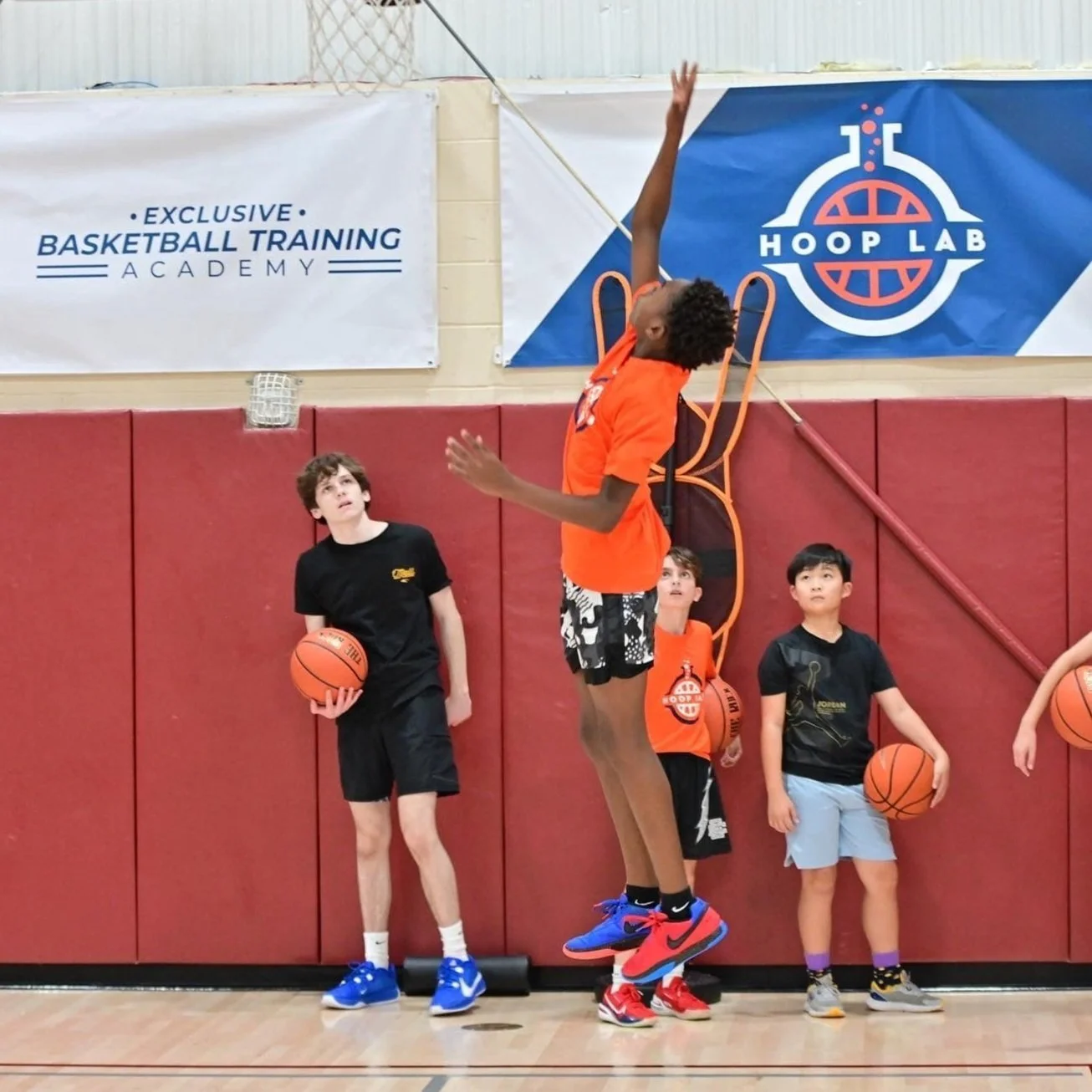 A young basketball player jumping in the air during practice at a sports facility, with three children standing nearby holding basketballs, against a red wall featuring signs for basketball training and Hoop Lab.