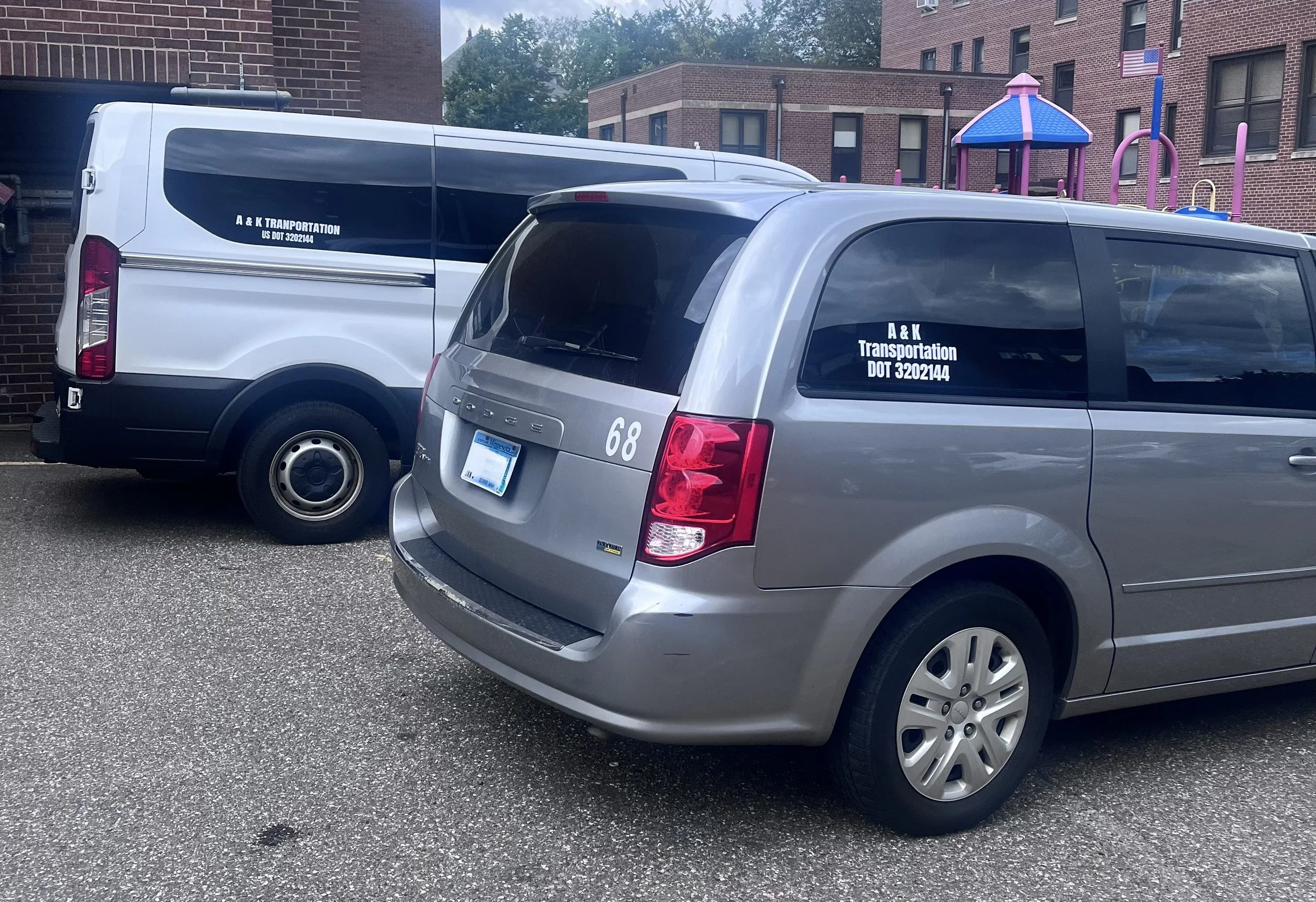 Silver Dodge van with the number 68 and black-tinted windows parked next to a white van with black accents, both marked with 'A & K Transportation DOT 3202144', in a parking lot near a brick building and colorful playground equipment.