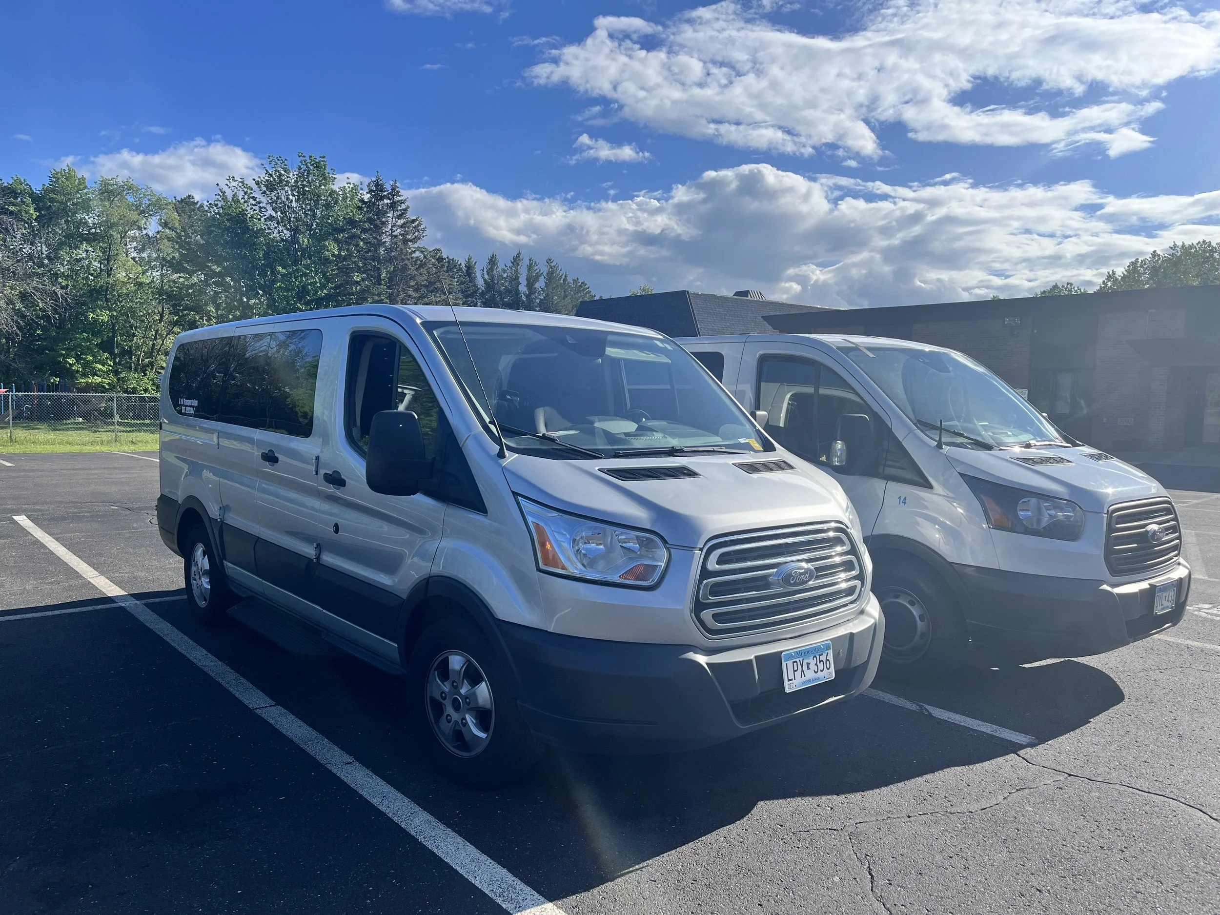 Two silver Ford Transit vans parked in a parking lot with a building and trees in the background, under a partly cloudy sky.
