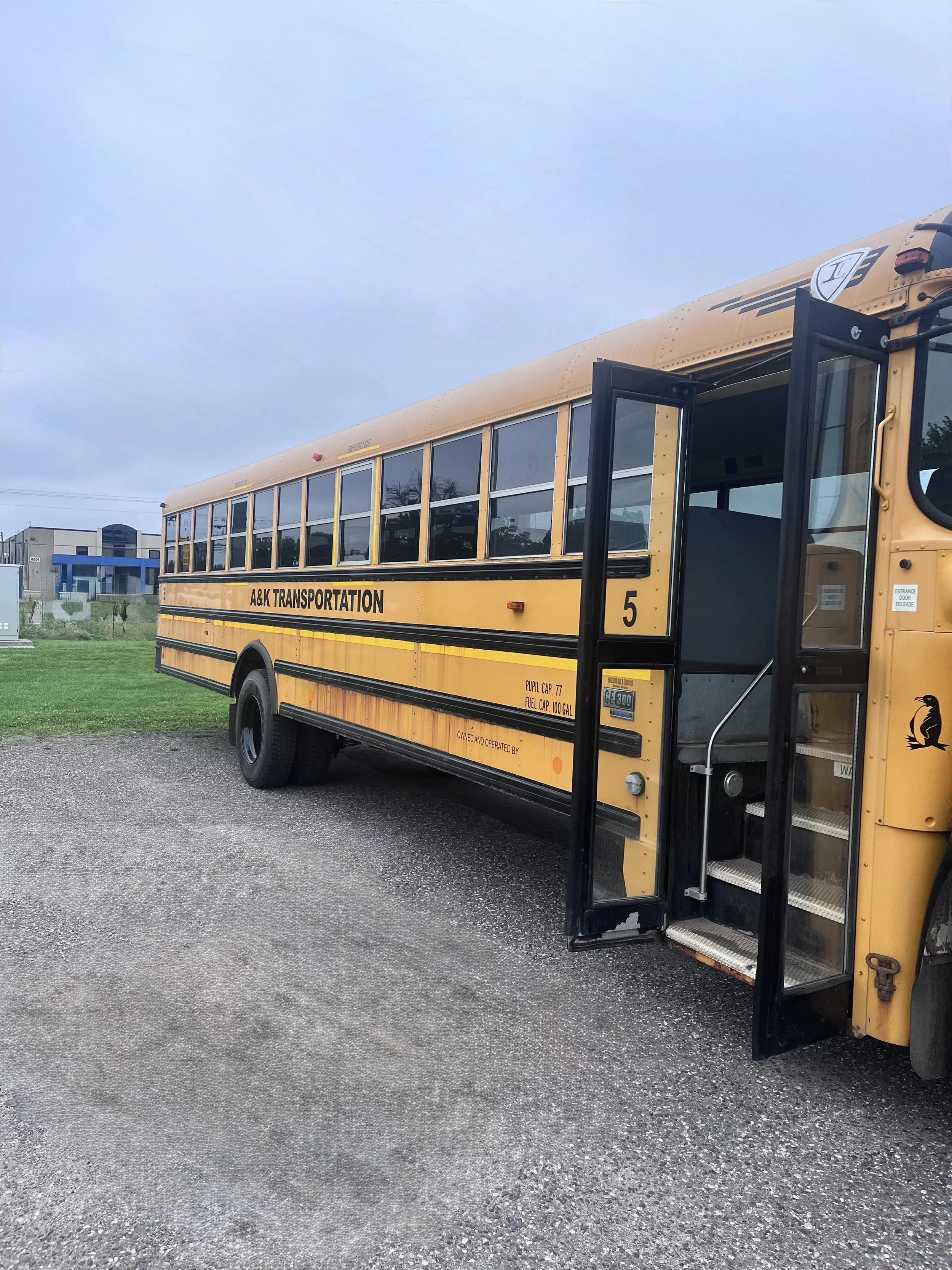 Yellow school bus with open door parked on gravel, with trees and a building in the background.