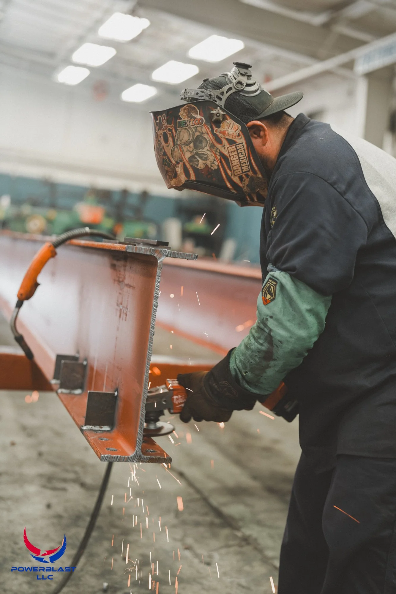 A worker wearing a decorated welding helmet, gloves, and protective clothing welding a metal structure in an industrial workshop with sparks flying.