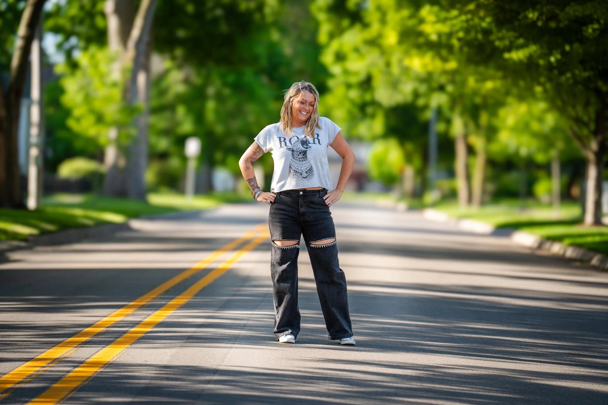 A woman with blonde hair, wearing a gray t-shirt with a dog graphic and the word 'ROAR,' standing in the middle of a street with green trees and yellow lines, smiling with hands on hips.
