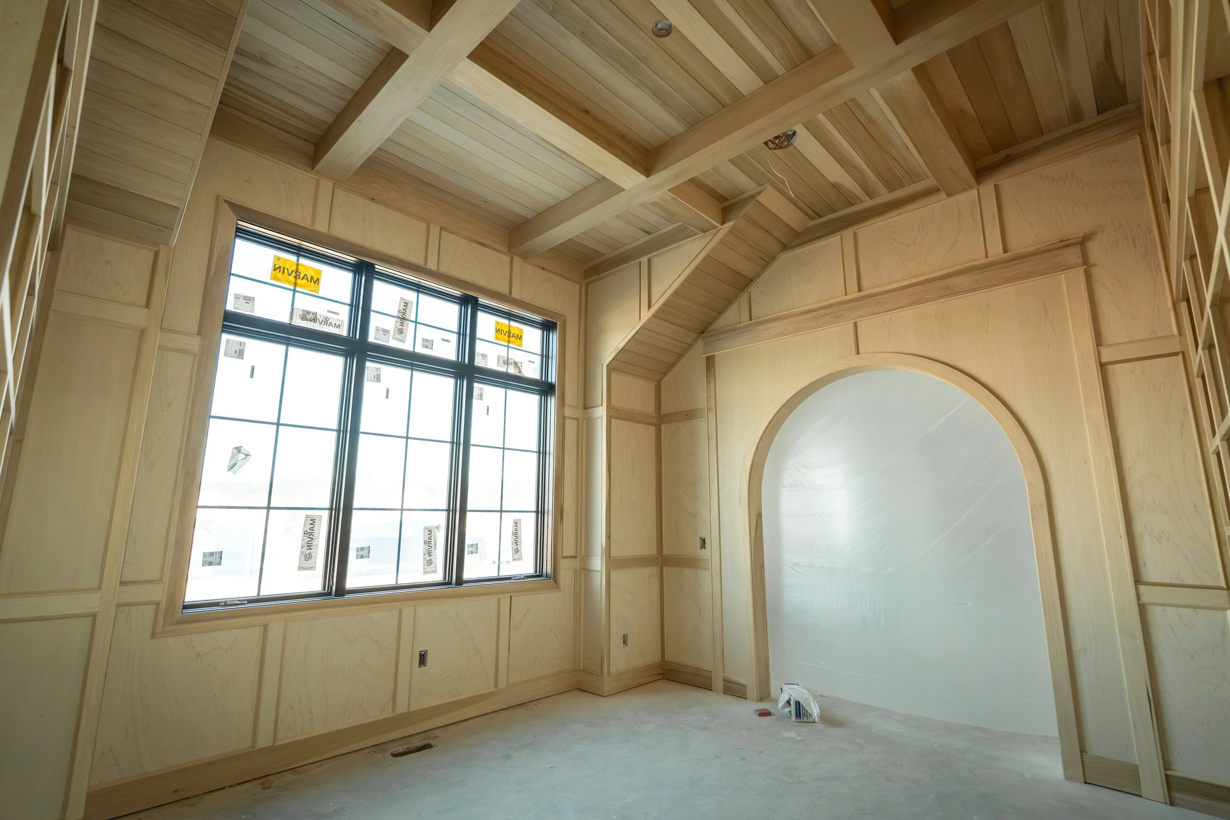 Interior view of a room under construction with light wood paneling on walls and ceiling, large window, and unfinished floor.