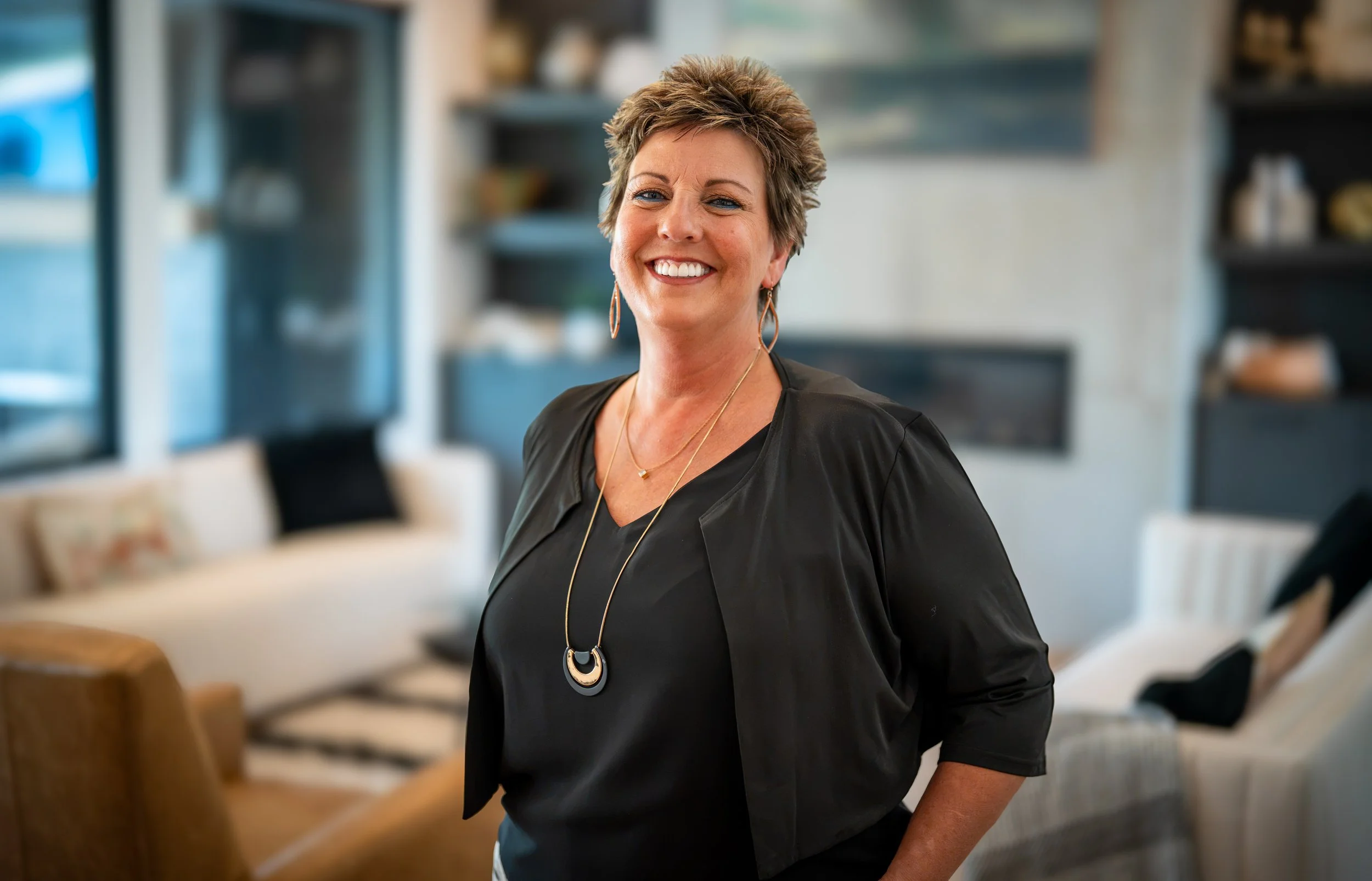 Smiling middle-aged woman with short, styled hair wearing a black top and jewelry standing in a modern, well-lit living room.
