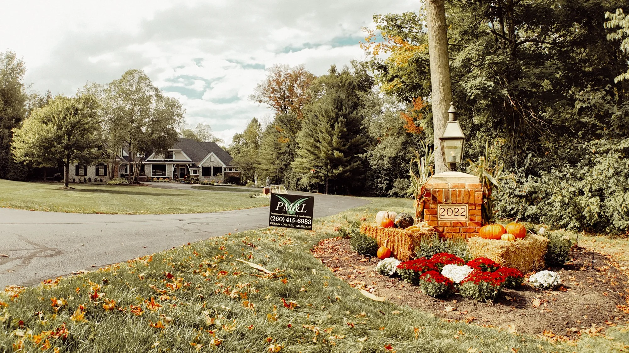 A suburban house with a manicured lawn and a fall-themed display near the driveway, including pumpkins, gourds, and flowers. There is a black sign for a landscaping business, and a brick mailbox decorated for autumn with a lantern and the year 2022. 