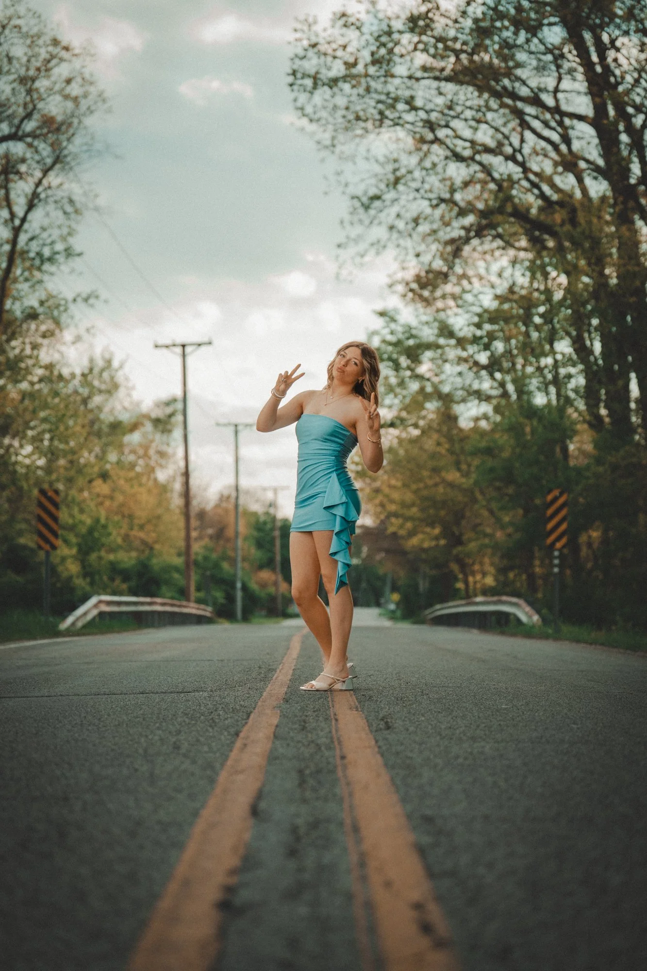 A young woman in a blue strapless dress and white high heels stands in the middle of a two-lane road, making a peace sign with both hands. The road is lined with trees and utility poles, with a partly cloudy sky above.