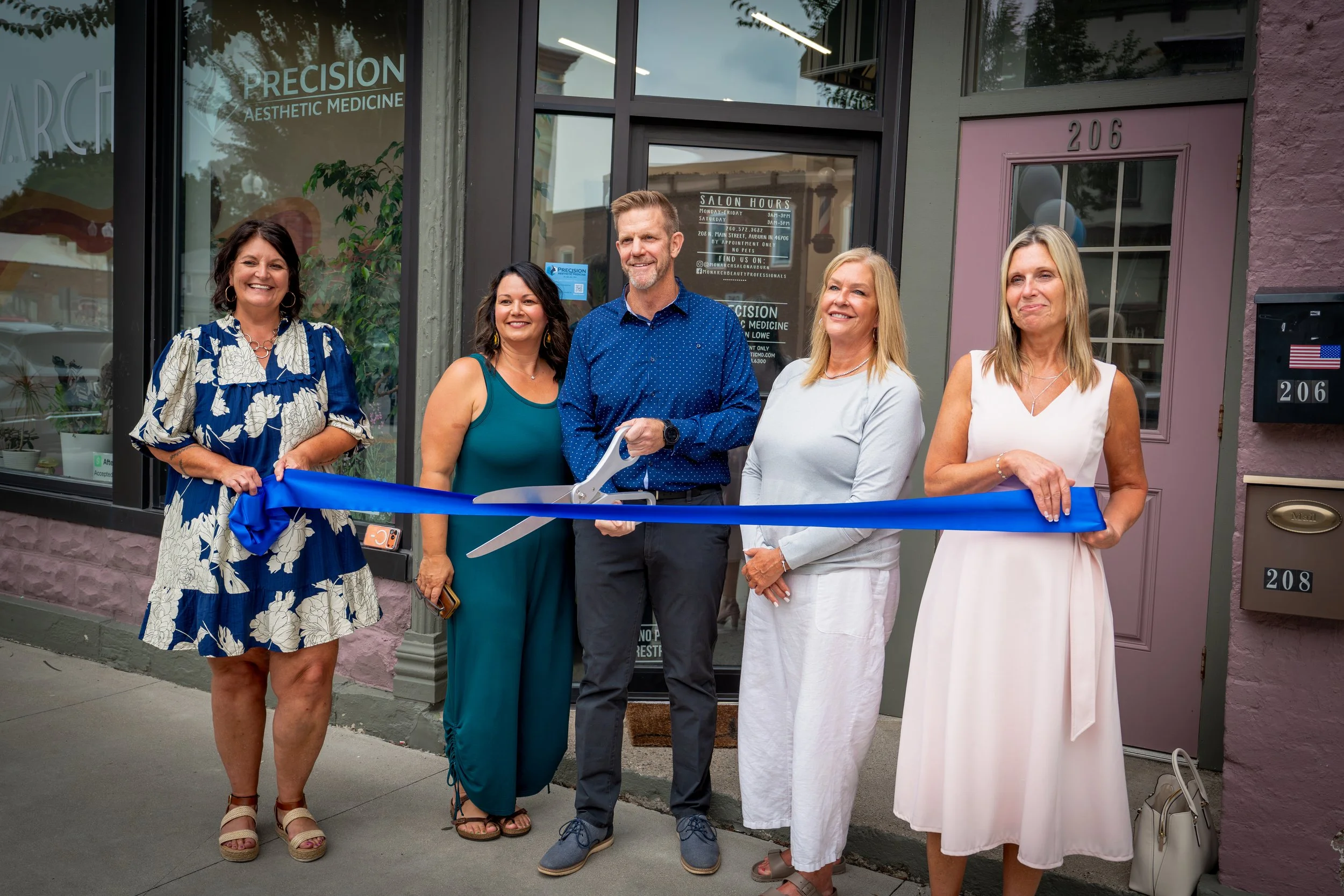 A group of five women and one man standing outside a storefront with a purple door, participating in a ribbon-cutting ceremony. The man is holding large scissors. The women are smiling and dressed in summer attire.