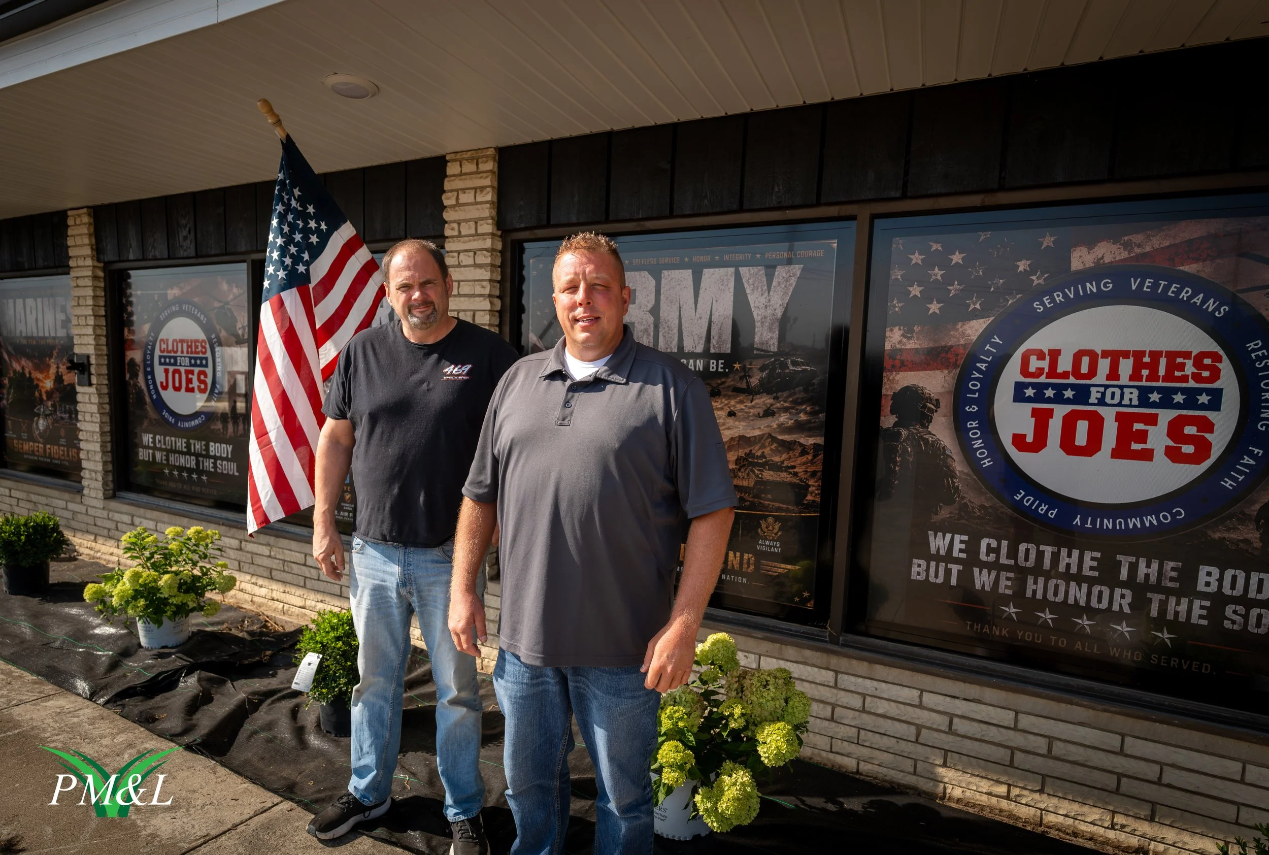 Two men standing outside a building with large window posters, one holding an American flag. The posters feature patriotic themes related to military service, veterans, and appreciation for their sacrifices.