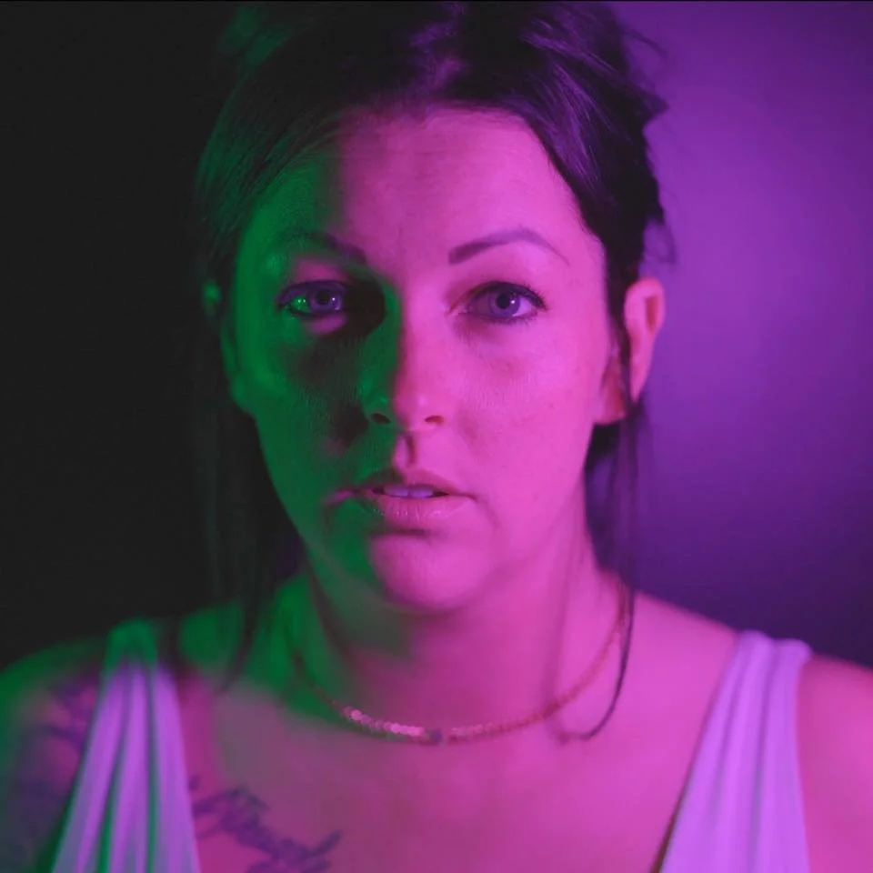 Portrait of a woman with short dark hair, illuminated by pink and purple neon lighting.