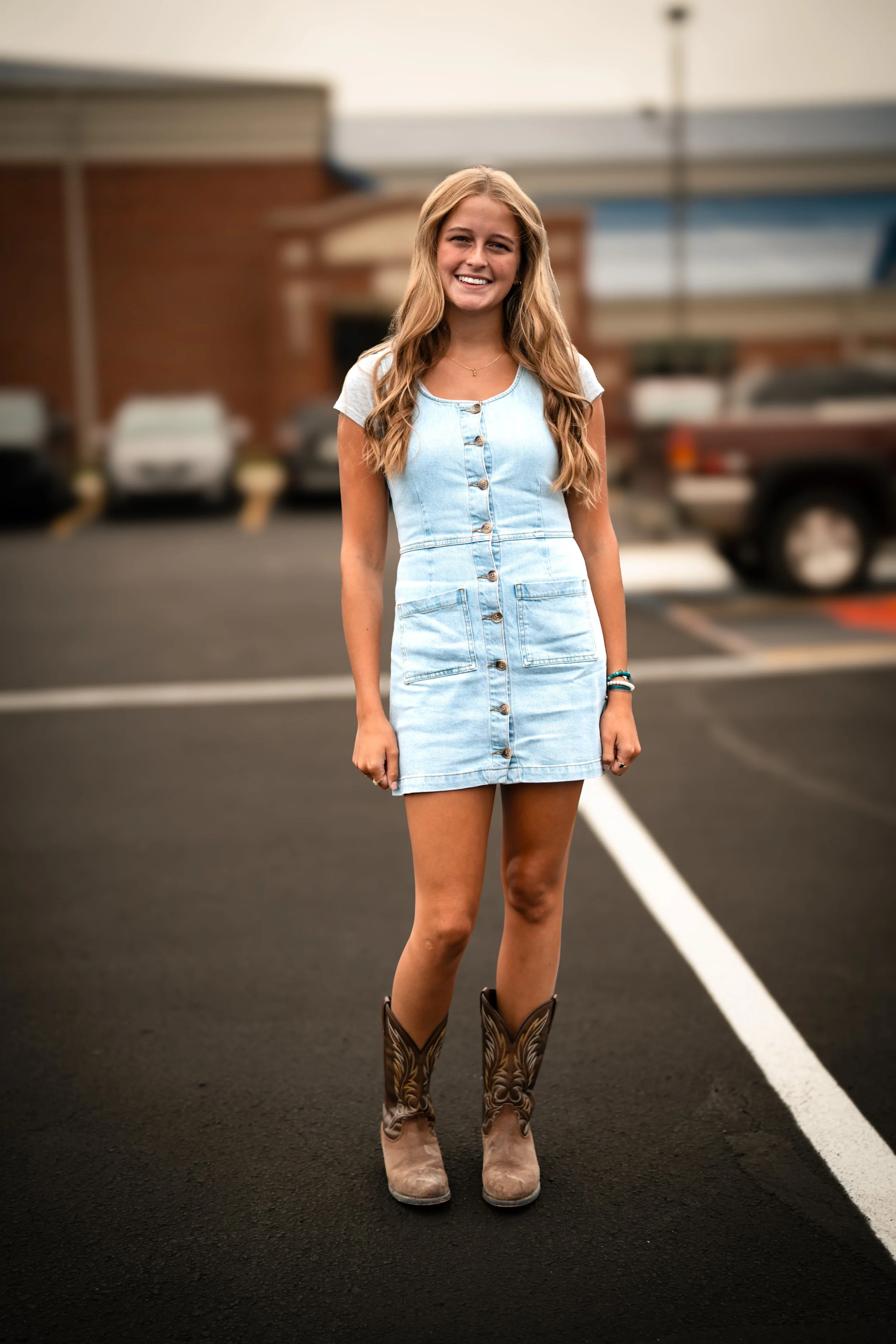 A young woman with long wavy blonde hair stands in the middle of a parking lot, smiling at the camera. She is wearing a light blue denim dress with buttons, cowboy boots, and bracelets on her wrist. The background shows parked cars and a building.