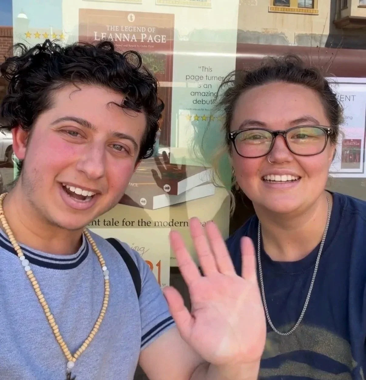 Addam Ledamyen and Theo Neeno smiling and waving at the camera, taking a selfie in front of a bookstore window that displays a poster about a book called "The Legend of Leanna Page."