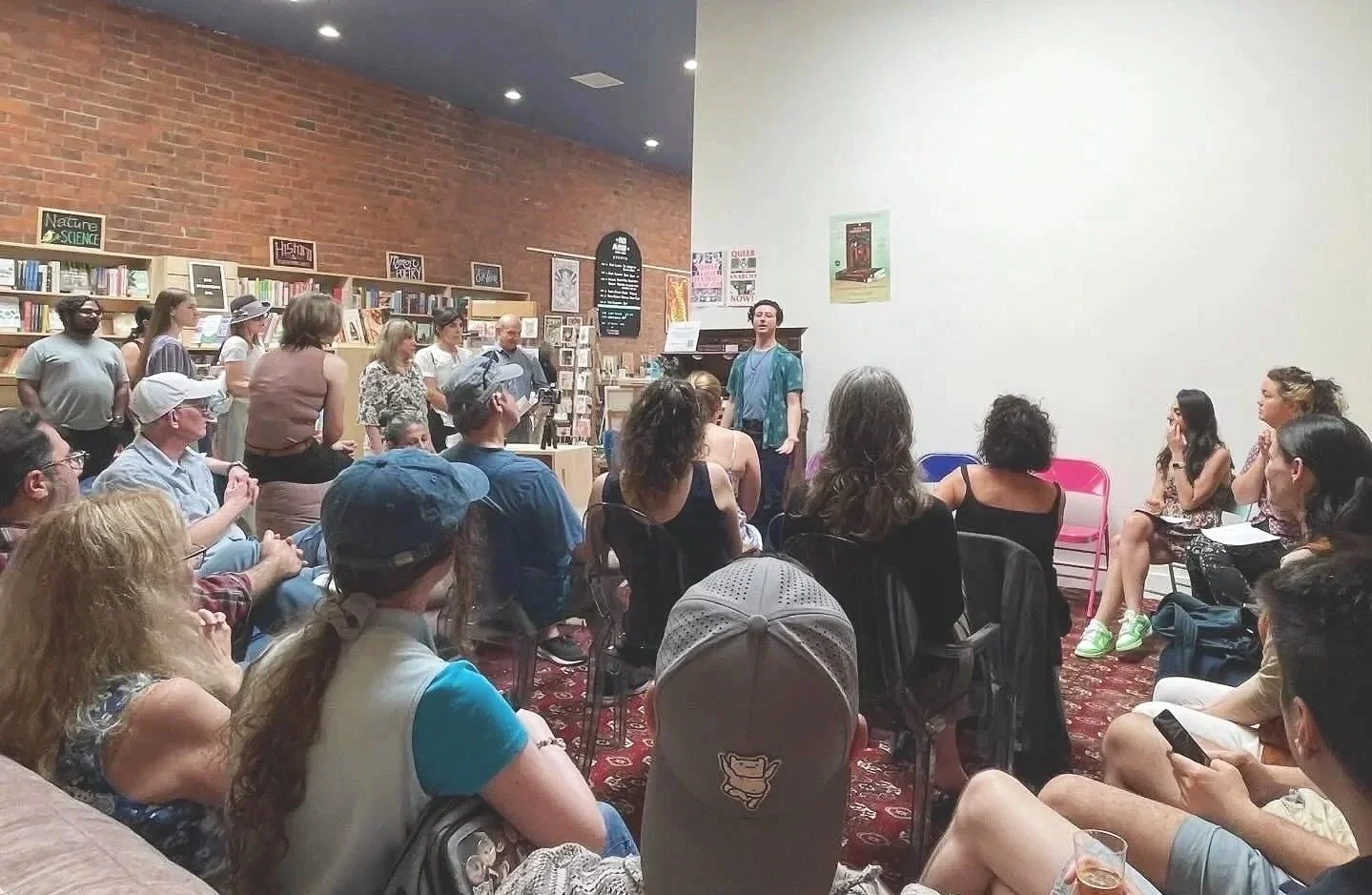 A person stands in front of an audience in a bookstore, with bookshelves and posters on the brick wall behind them, while the audience listens attentively.