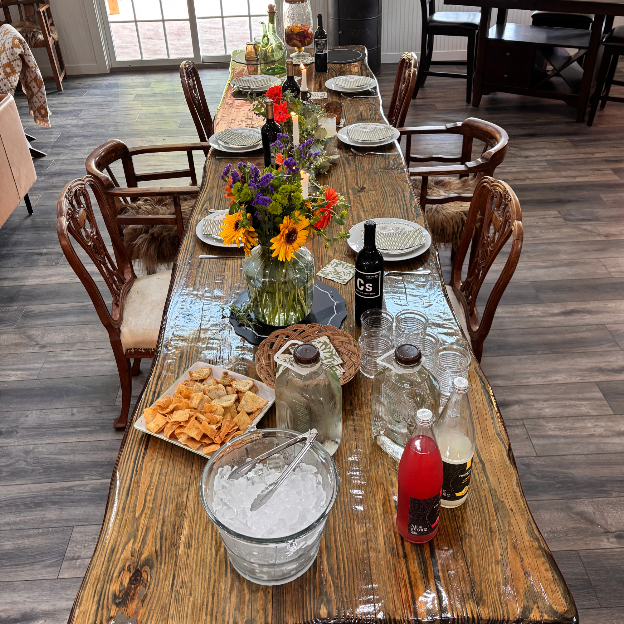 A wooden dining table set with plates, napkins, wine bottles, glasses, candles, and a colorful flower centerpiece, with chairs around it in a room with a sliding glass door in the background.