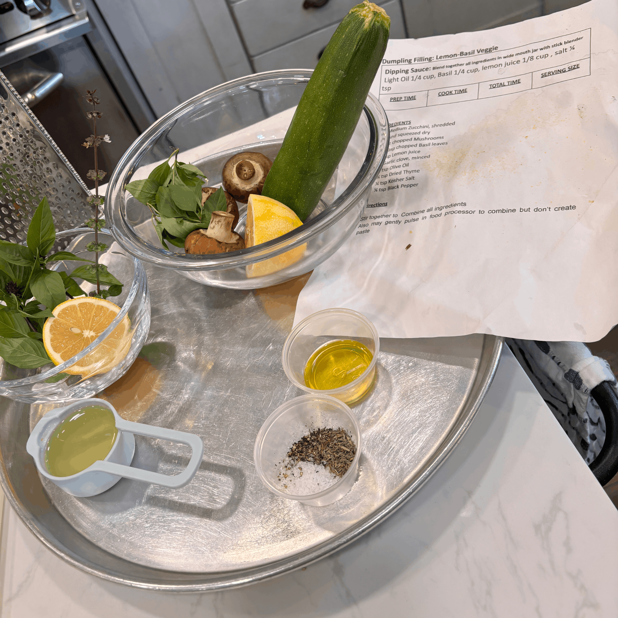 A glass bowl filled with fresh herbs, a lemon wedge, mushrooms, and chopped basil, placed on a kitchen countertop alongside a recipe sheet for lemon-basil veggie dumplings, with two small containers of sauce or oil nearby.