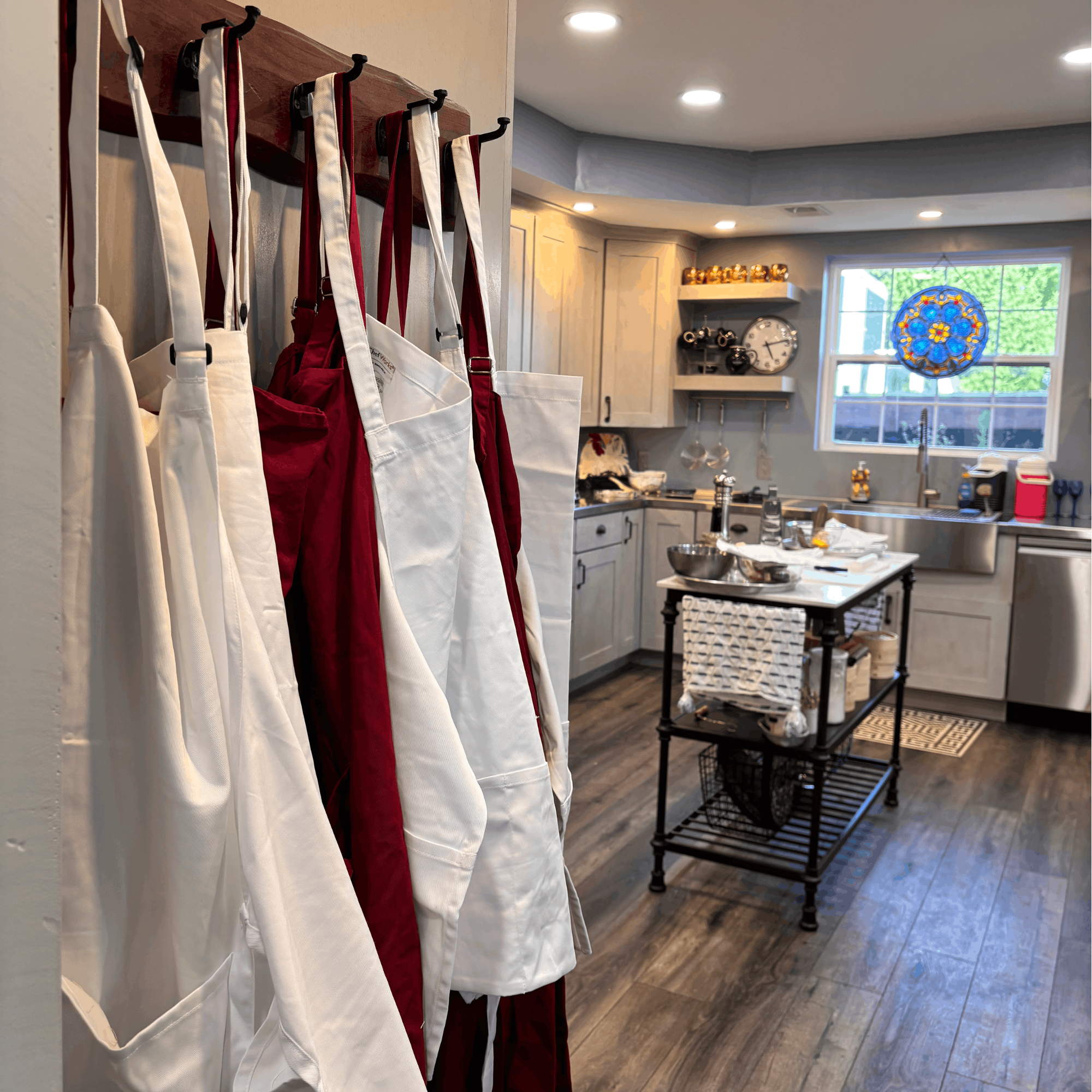 Kitchen with beige cabinets and a window with stained glass design, black shelves holding pots, and a black kitchen island with a large mixing bowl on top. Red and white aprons hanging on hooks in the foreground.