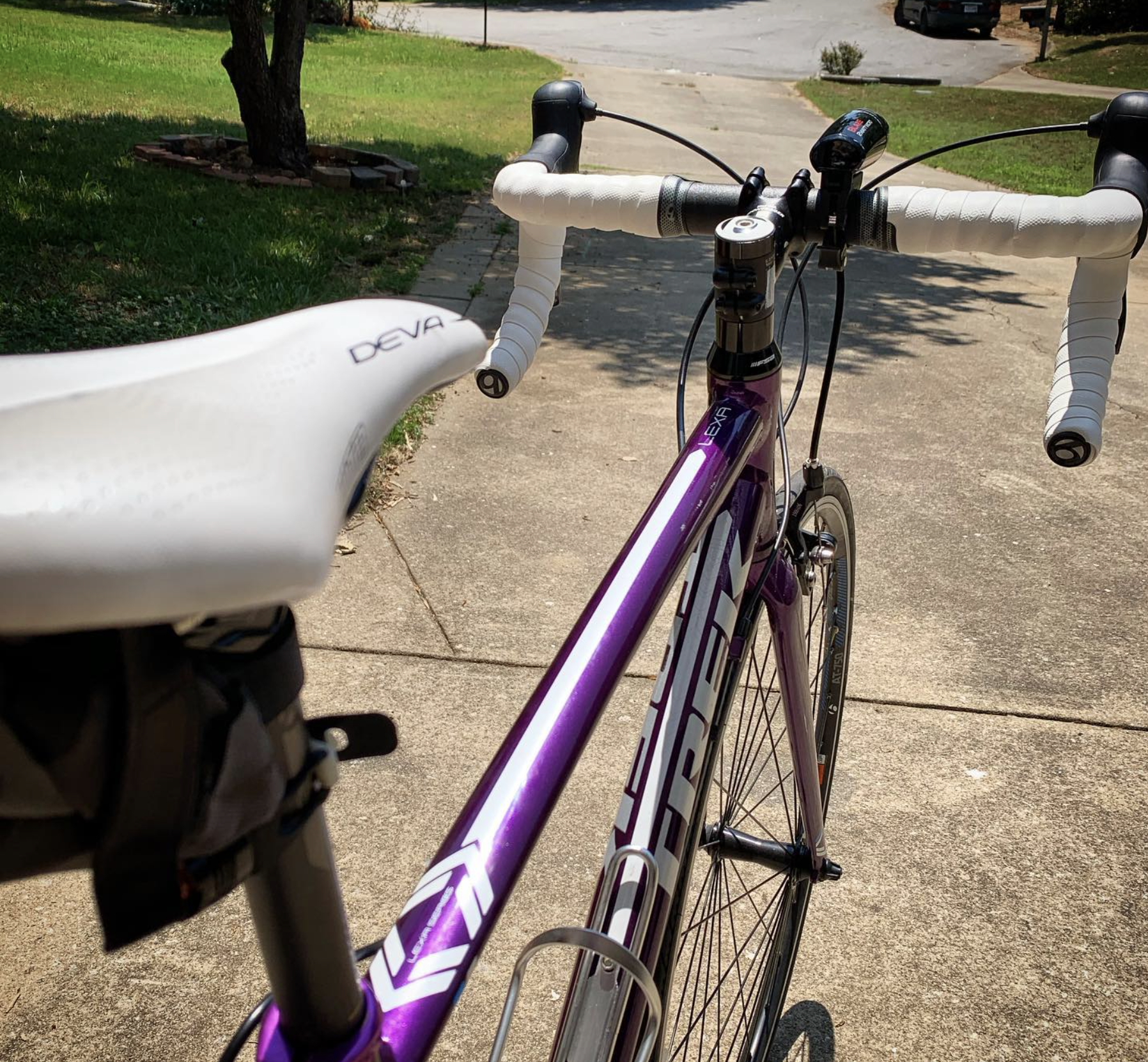A purple Trek road bicycle on a concrete sidewalk in sunlight, with white handlebar tape, a white saddle, and a small black pouch attached to the front of the saddle, surrounded by green grass, a tree, and a parking lot in the background.