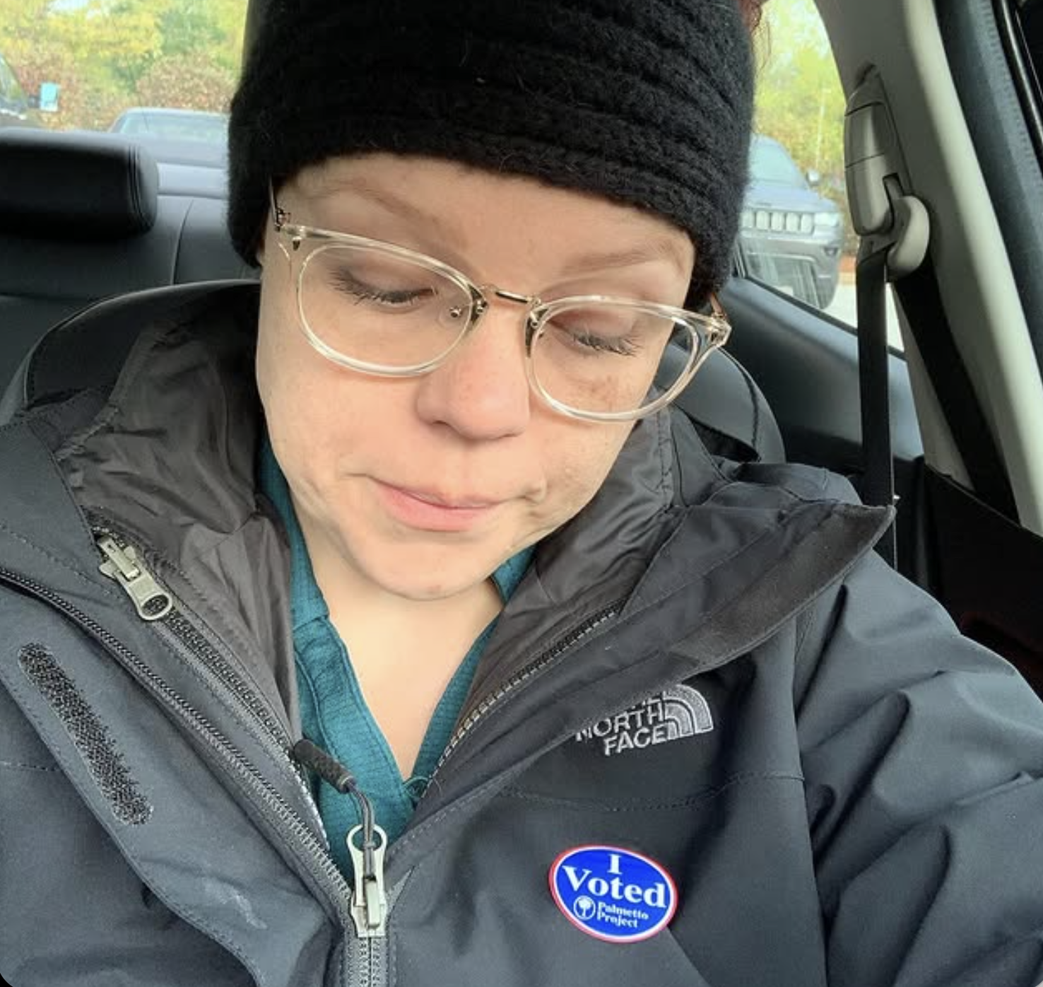 Person wearing glasses, a black beanie, and a North Face jacket with an "I Voted" sticker on it, sitting inside a vehicle.