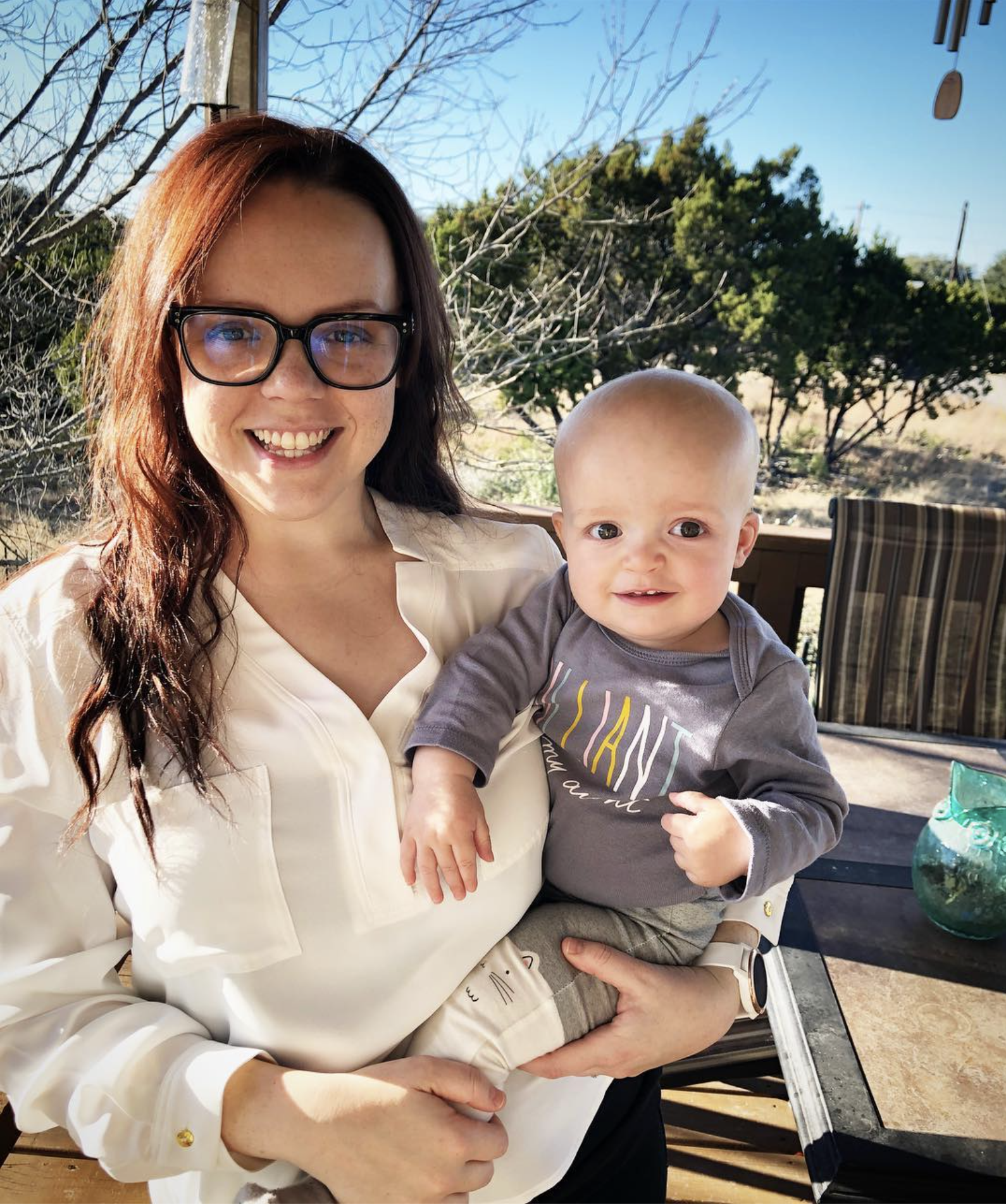 A woman with glasses and a big smile holding a toddler outside on a sunny day.