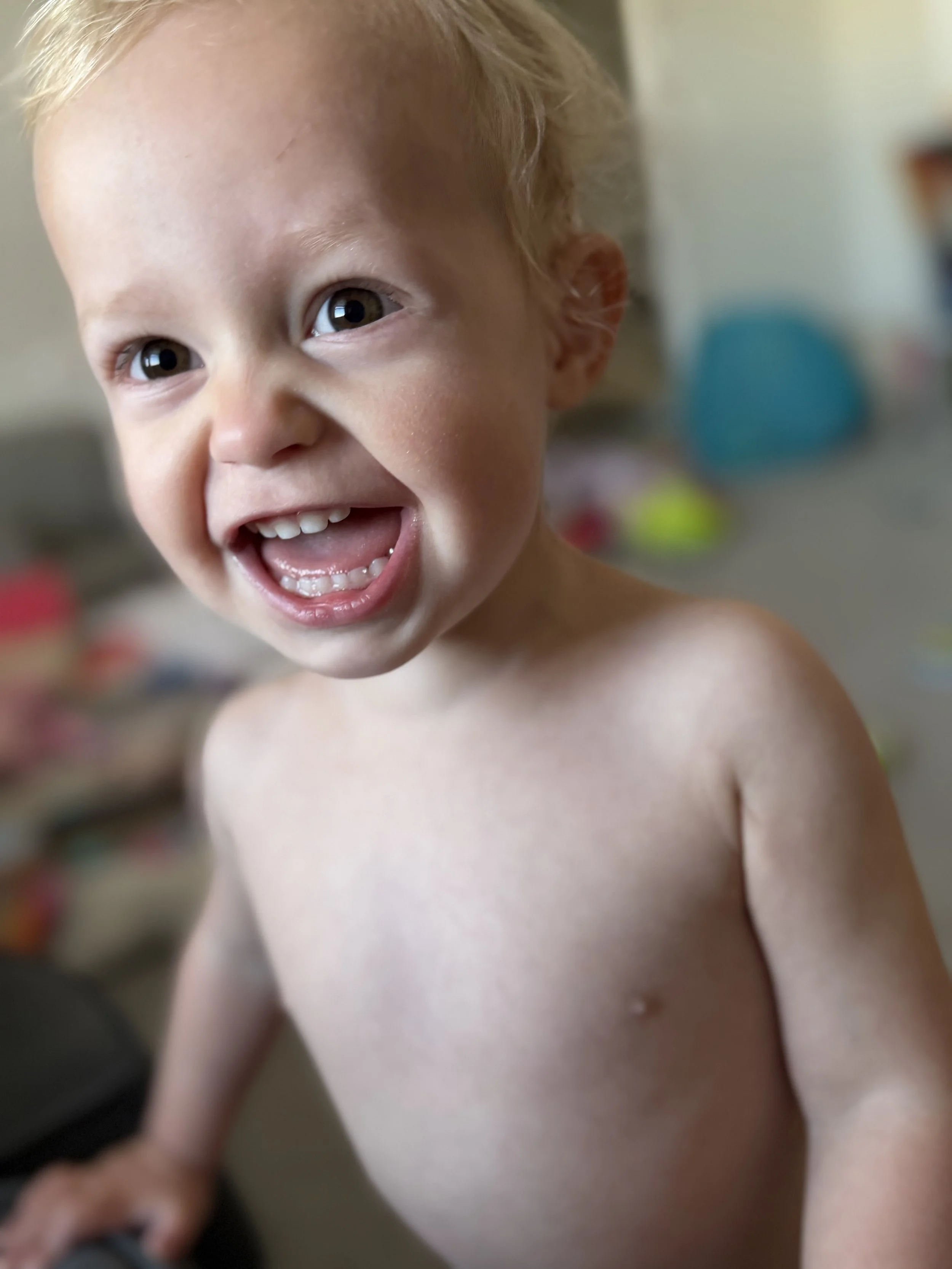 A young boy with blonde hair and brown eyes smiling, showing his teeth, and shirtless indoors with blurred background.
