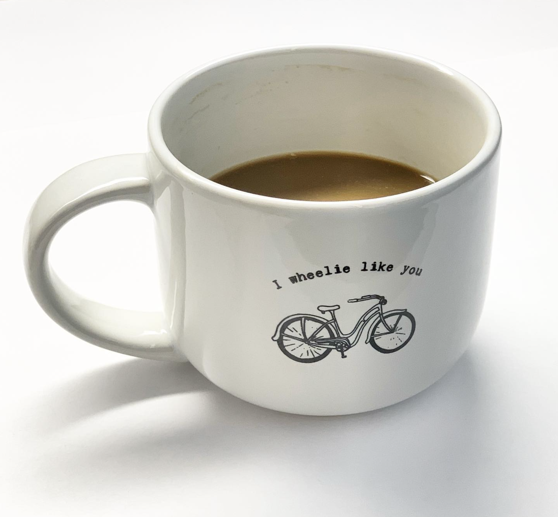 A white mug with a bicycle illustration and the text "I wheelie like you" filled with coffee on a white background.
