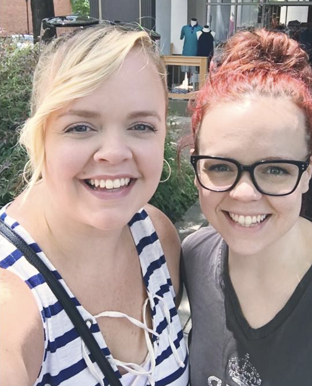 Two smiling women taking a selfie outdoors with store window and mannequins in the background.