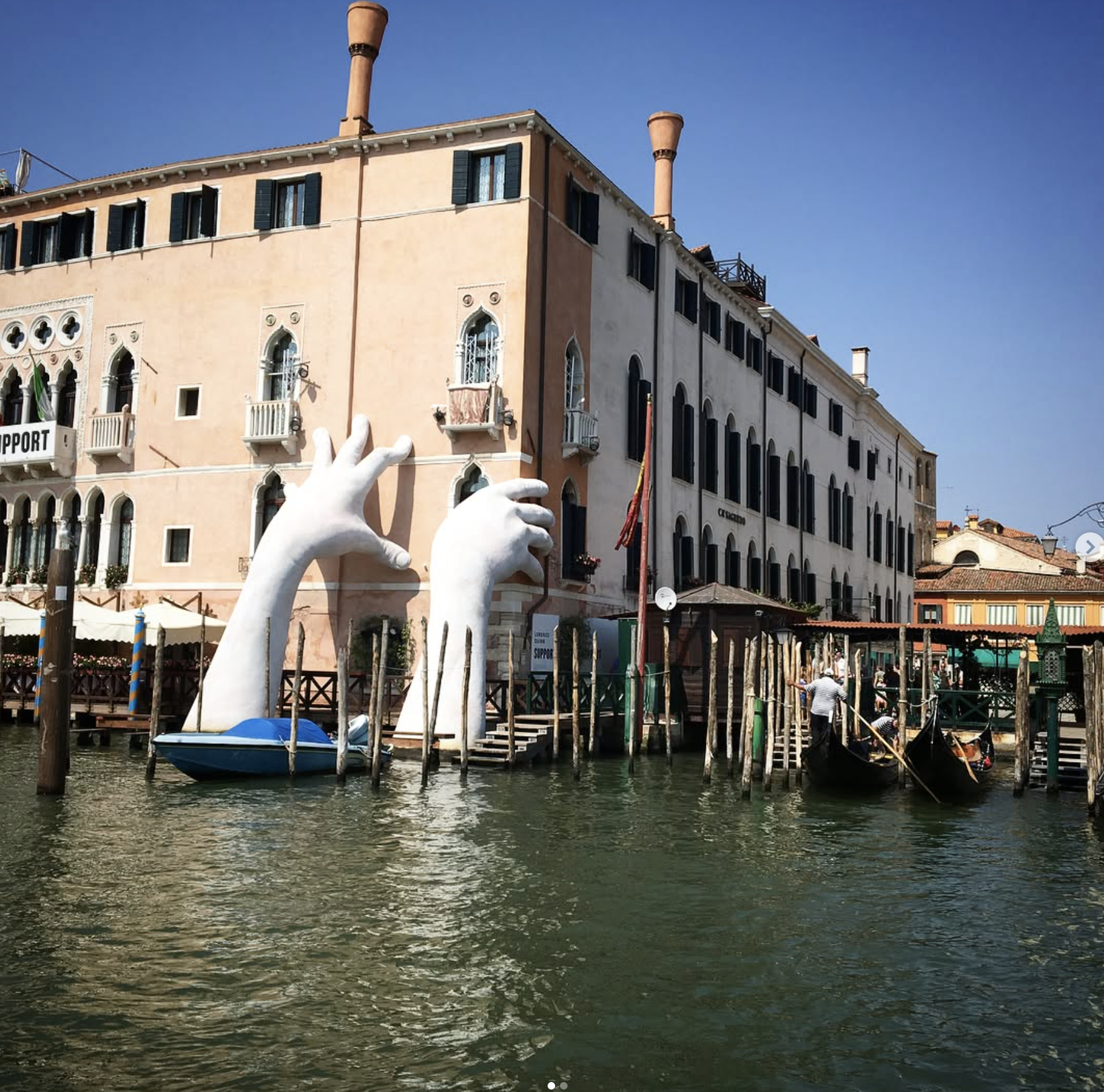 Large white sculpture of two hands emerging from a canal, with a small boat beneath them, located in front of a European-style building with arched windows and shutters along the water.
