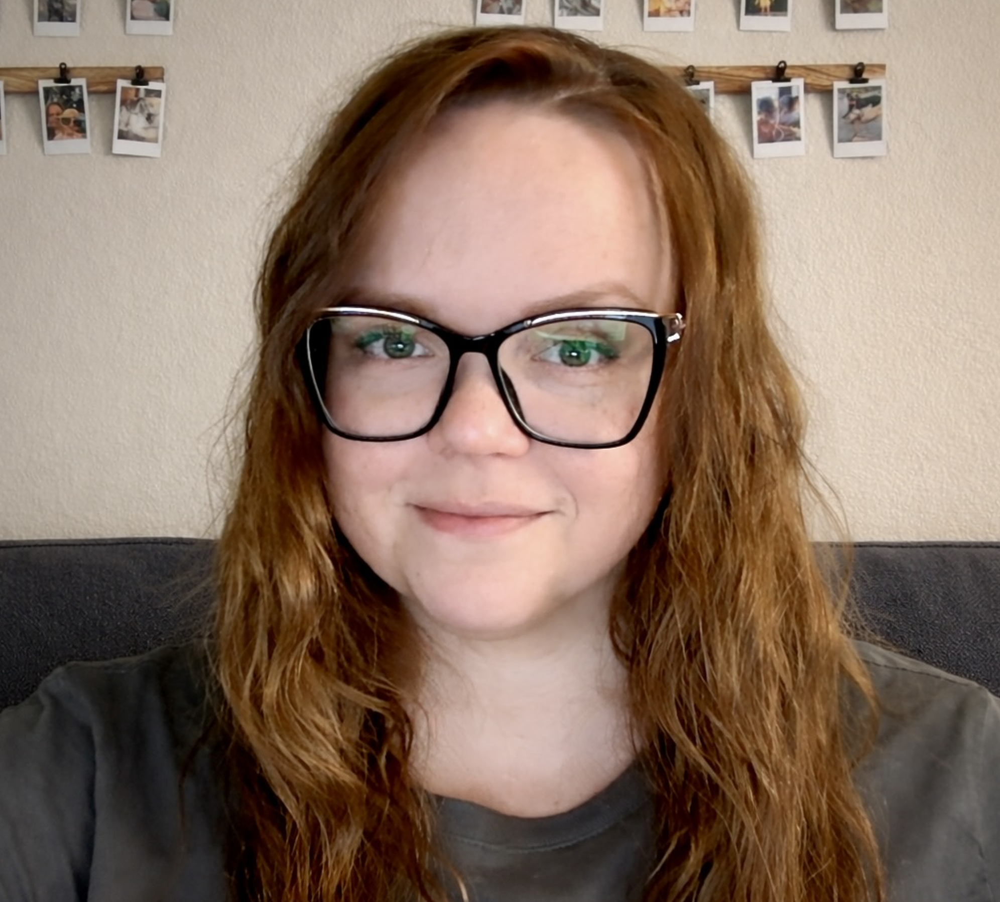 A woman with curly red hair wearing glasses, sitting on a couch with a wall behind her decorated with photographs hanging on a string.