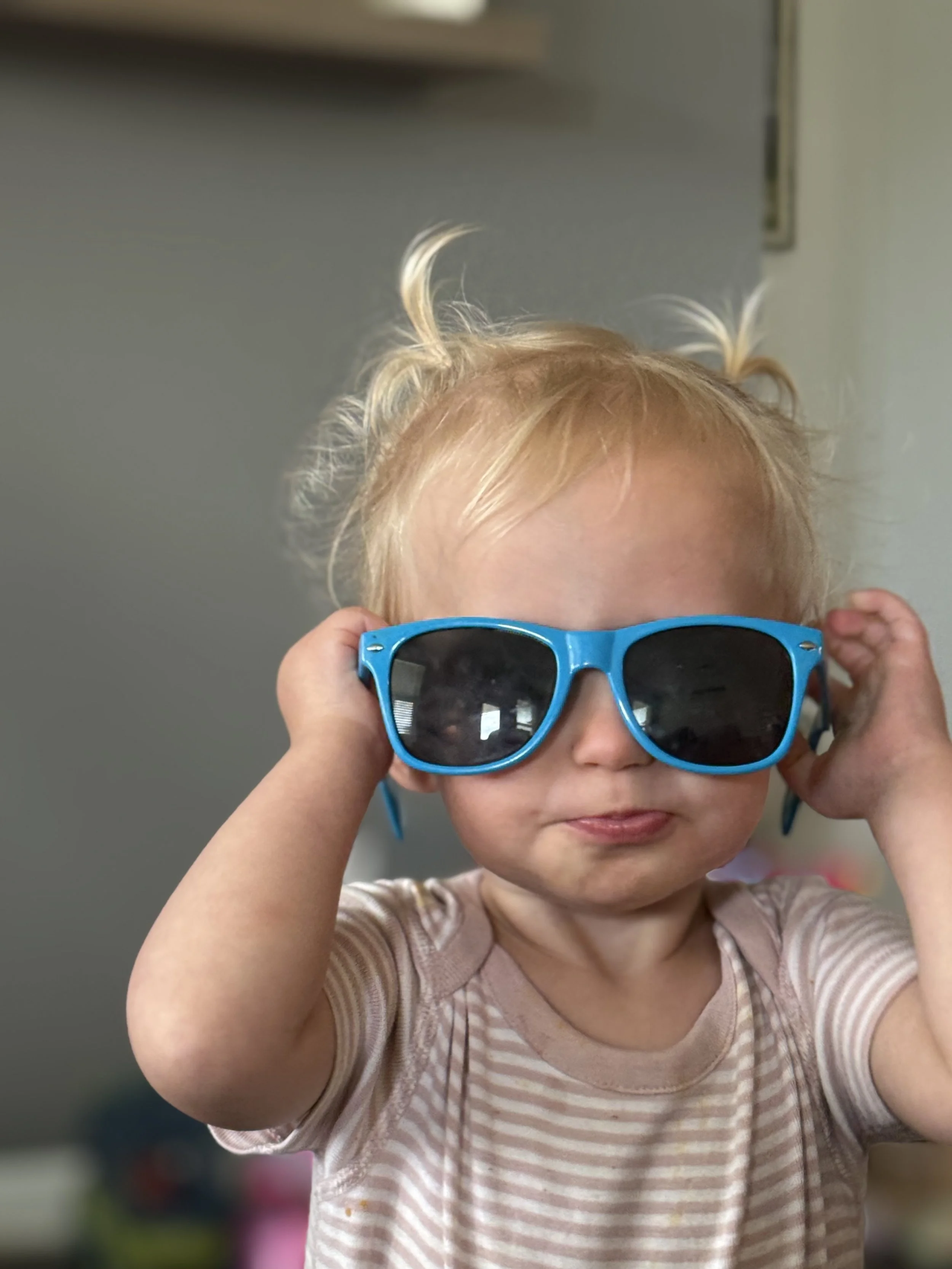 A young child with blonde hair in pigtails wearing blue sunglasses and a beige striped shirt indoors.