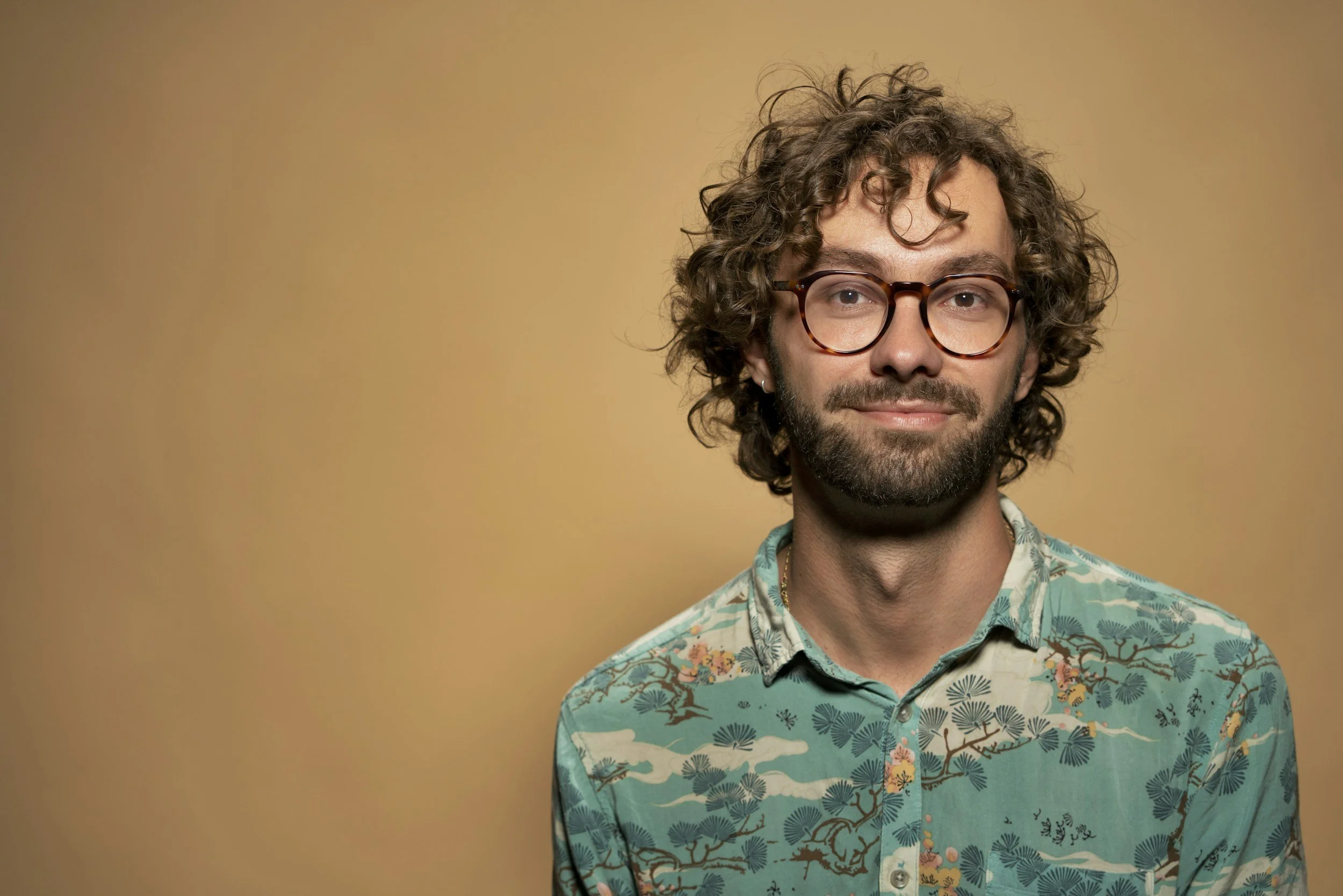 A man with curly brown hair, glasses, a beard, and earrings wearing a patterned shirt, standing against a plain beige background.