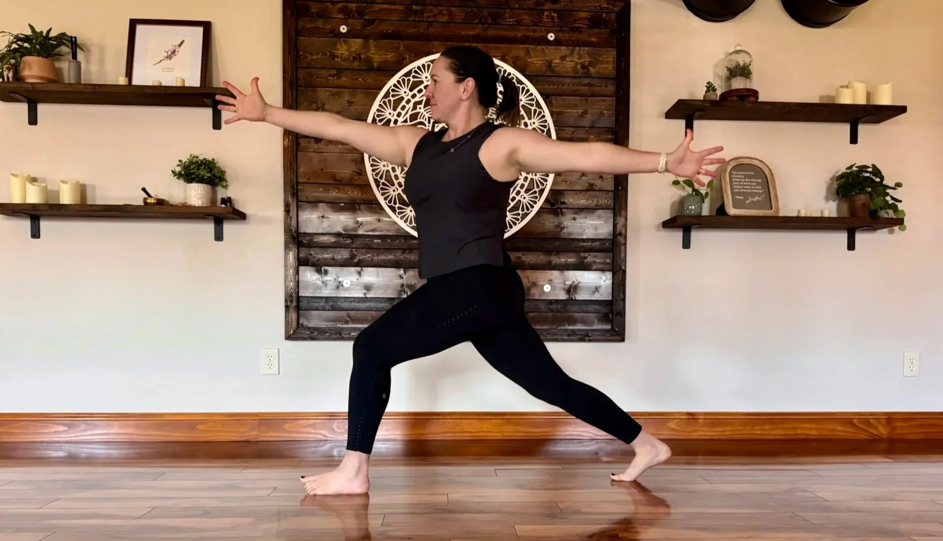 A woman practicing yoga indoors, in a warrior pose, with arms extended and one knee bent, on a wooden floor in front of a wall with floating shelves, plants, and decorative items.