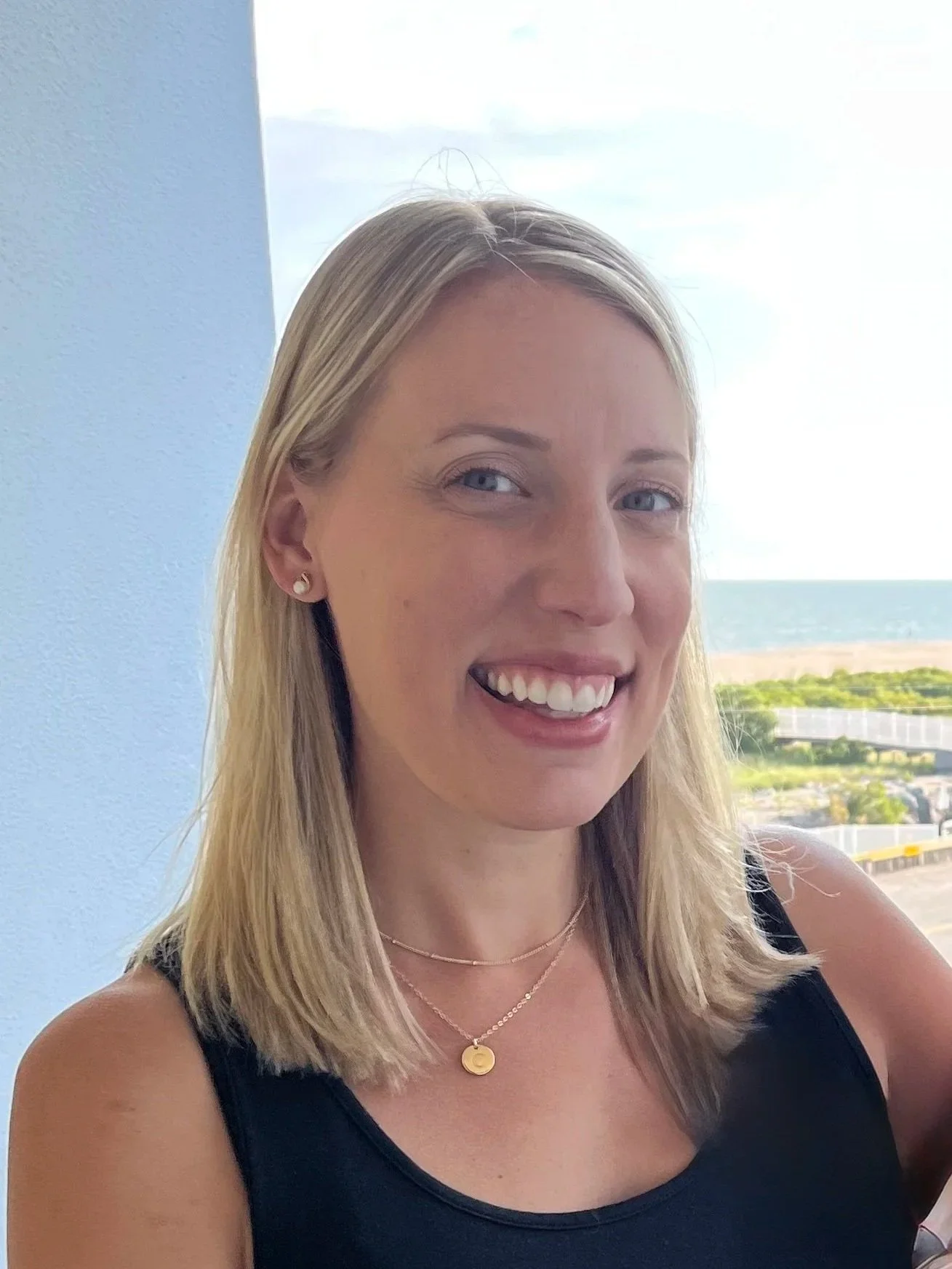 A smiling blonde woman wearing a black sleeveless top and gold jewelry, standing by a window with an ocean view in the background.