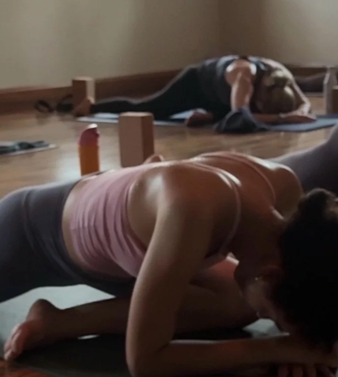 Two women practicing yoga in a dimly lit studio, one on her knees with her head lowered, and another lying face down on a mat in a child's pose.