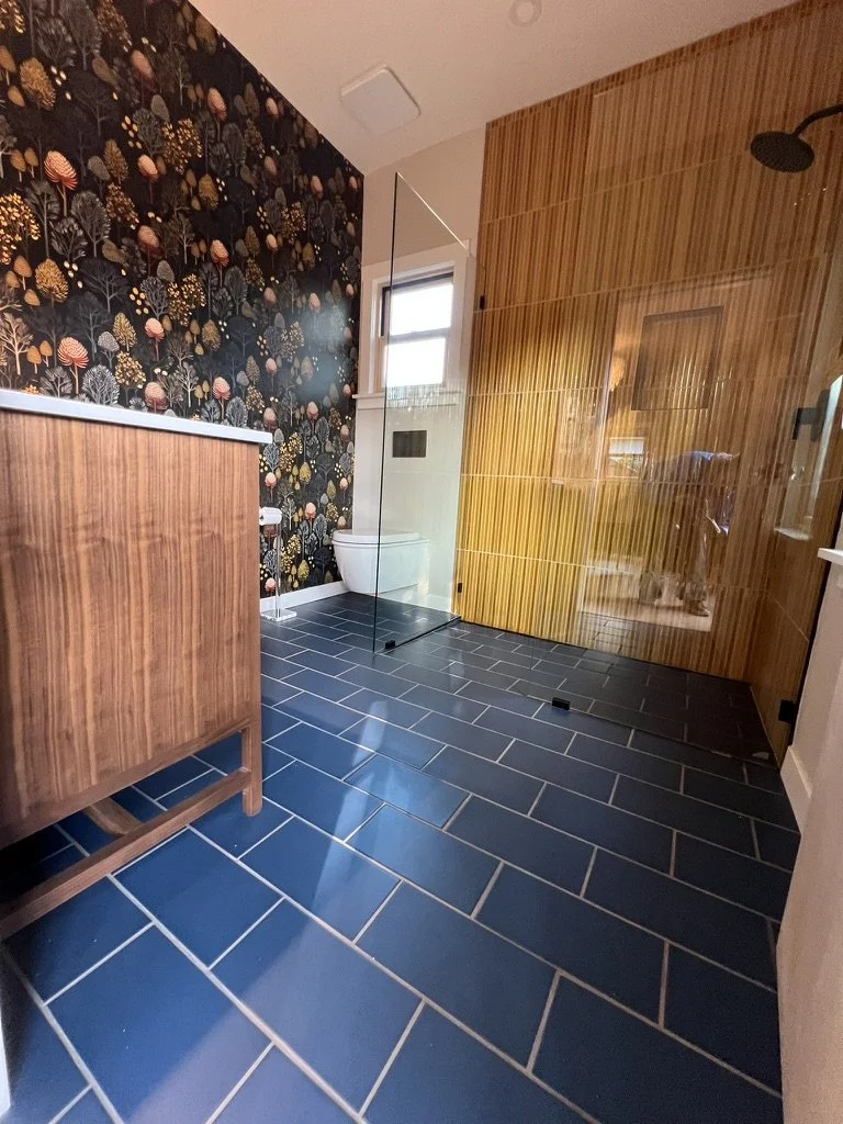 Modern bathroom with black and gold floral wallpaper, wooden cabinetry, blue tiled floor, a toilet, and a shower area with a glass partition and yellow tiles.
