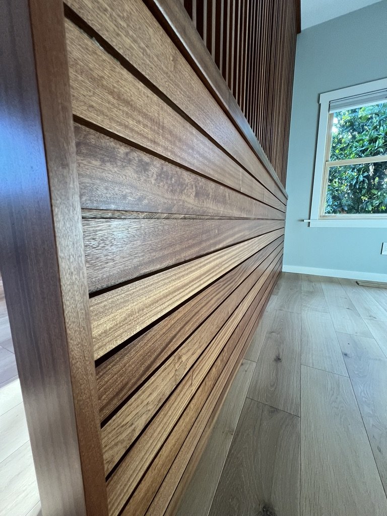 Close-up of a wooden slatted wall and wooden floor in a room with a window showing greenery outside.