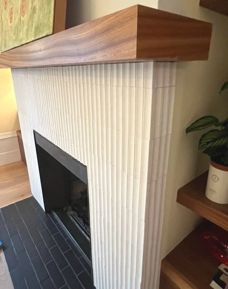 Close-up of a fireplace with a beige, textured tile surround, topped with a wooden mantel, and dark brick hearth.