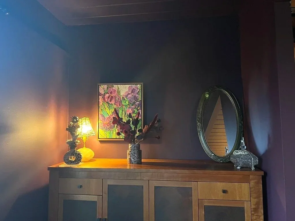 A wooden sideboard against a dark blue wall decorated with a framed floral painting, a vase with dark flowers, a lit yellow lamp, a decorative glass container, and an oval mirror.