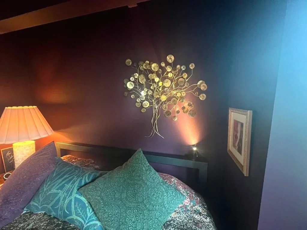 A bedroom with dark walls, a gold-colored wall art resembling a tree with circular leaves, a framed picture, a bedside lamp with a beige shade, and pillows on the bed with patterned and textured covers.