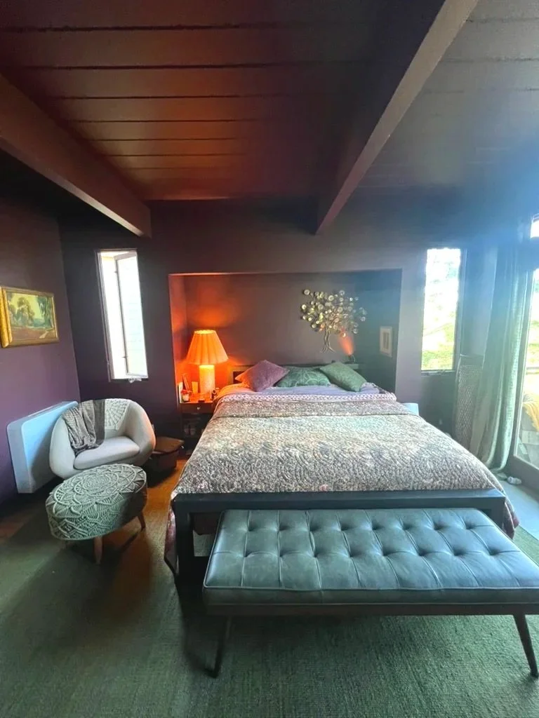 A cozy bedroom with a sloped wooden ceiling, a large bed with green and purple pillows, a bedside lamp emitting warm light, a small loveseat with a blanket, an ottoman, and large windows letting in natural light.