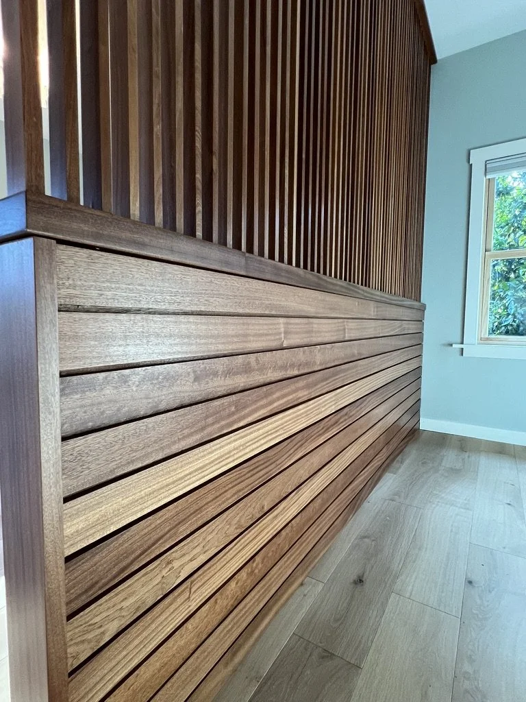 Close-up of a wooden slatted wall with varying shades of brown, next to a window with greenery outside.