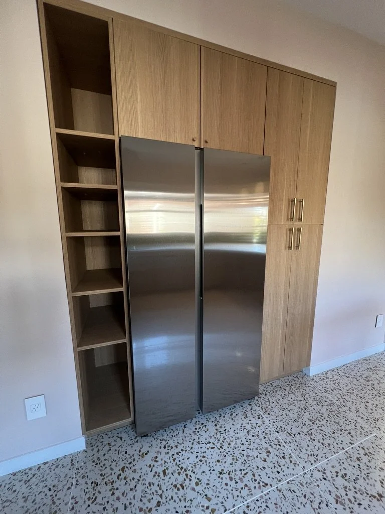 Kitchen cabinet with wood finish and open shelving on the left, stainless steel double-door refrigerator in the center, and additional wood cabinets with gold handles on the right. The floor has a speckled terrazzo pattern.
