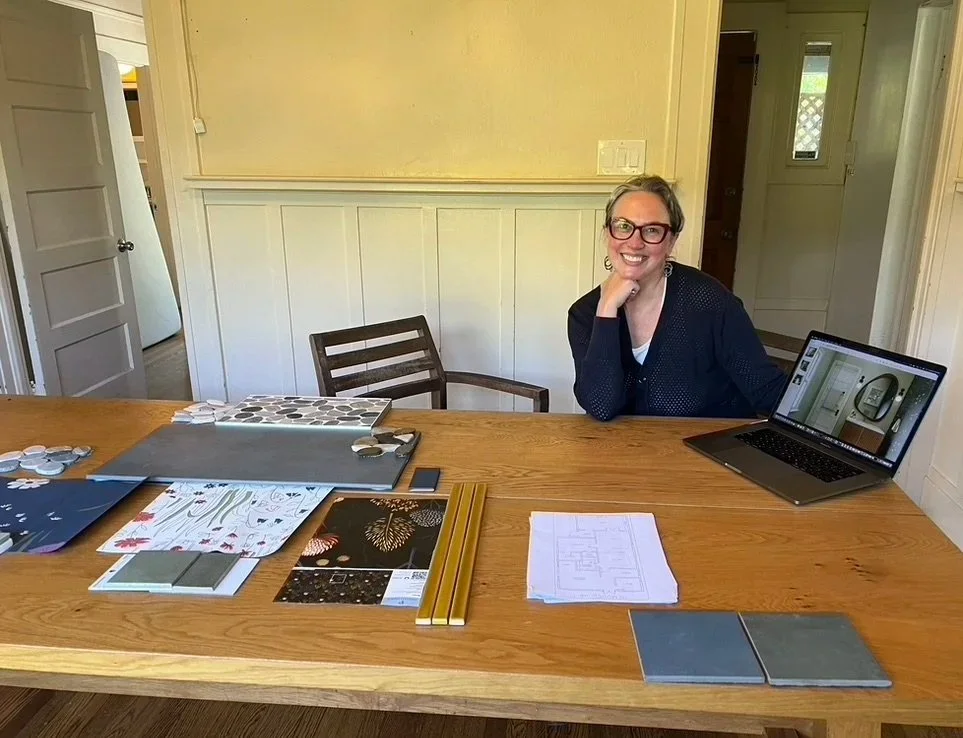 Woman sitting at a wooden table with design tiles, samples, and a laptop in a home interior.