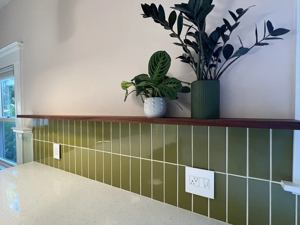 Two potted plants on a wooden ledge above a kitchen counter with a green tiled backsplash and white outlets.