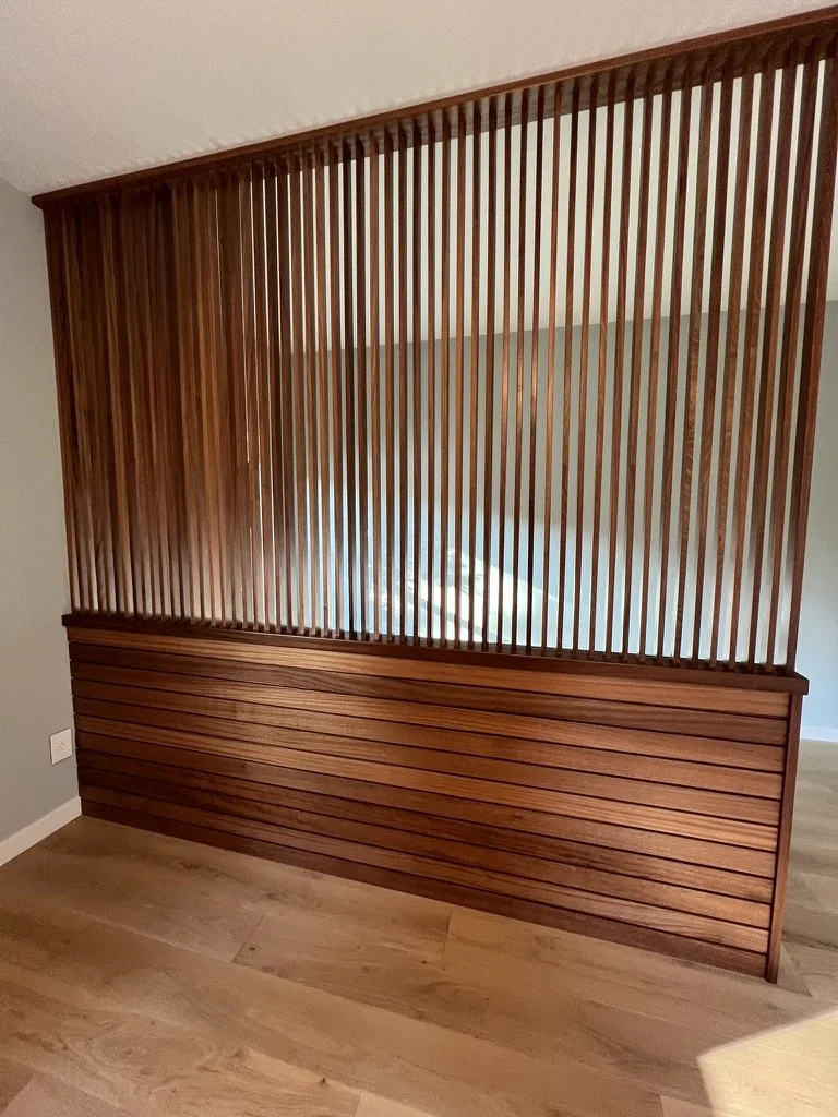 Wooden room divider with vertical slats and horizontal panel at the bottom, placed against a wall with floor.