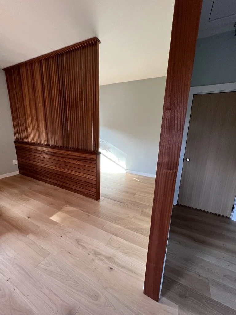 Split-level interior space with light wooden flooring, a wooden paneled wall, and beige walls with a stairway opening in the background.