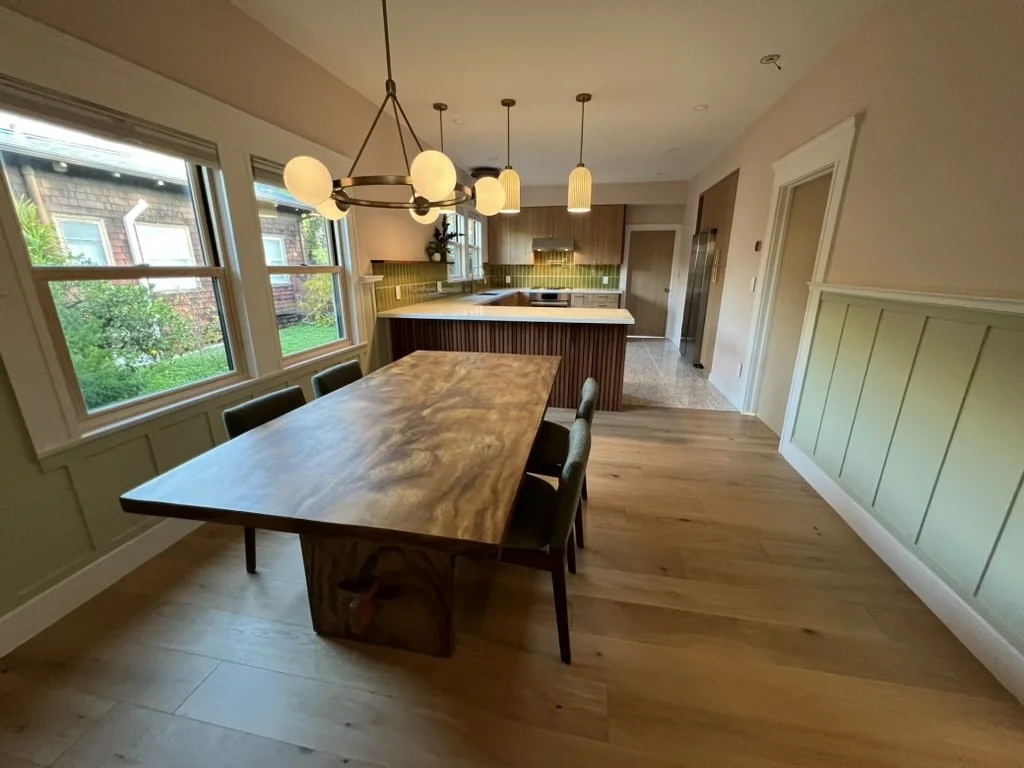 Dining area with wooden table and six chairs, large windows showing a backyard, and a kitchen with green backsplash and hanging pendant lights.