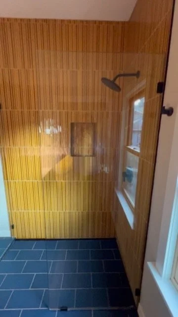 A shower area with wooden paneling, a ceiling-mounted showerhead, a small shelf, and a window on the door.