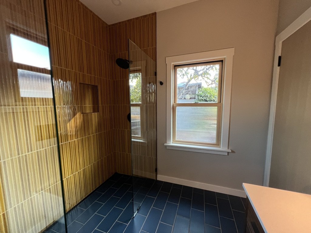 A bathroom with a walk-in shower that has glass walls and yellowish-brown tiles, a window with a view of trees outside, and a small part of a countertop visible.