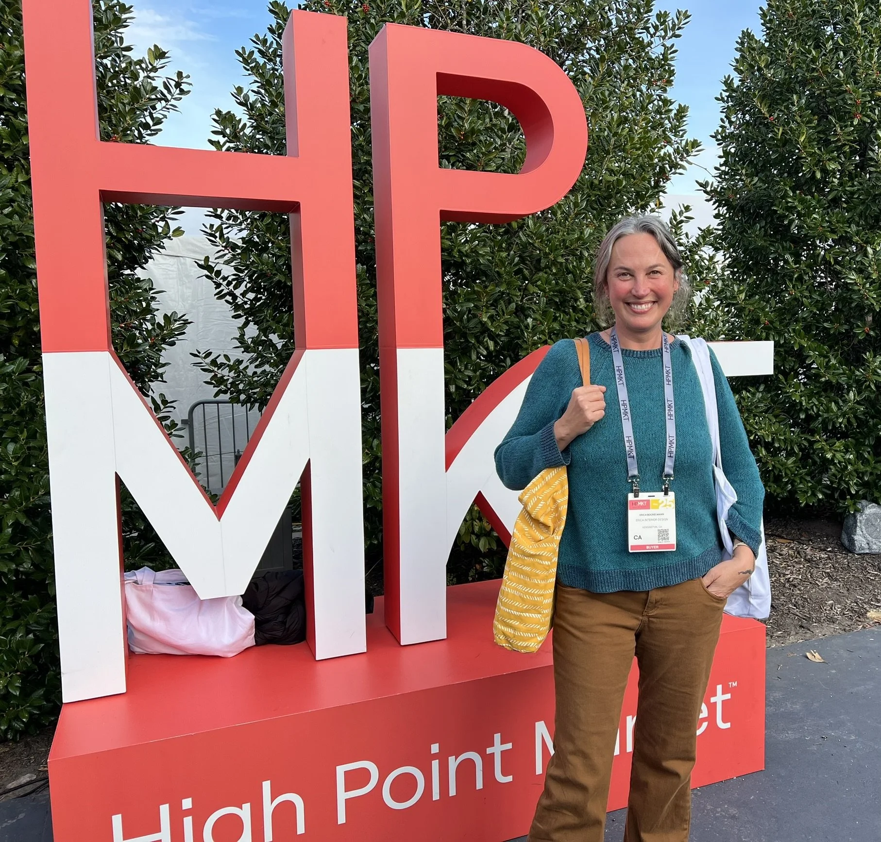 A woman standing in front of a large sign that reads 'HPM' at the High Point Market. She is smiling, carrying a yellow bag, and wearing a blue-green sweater, brown pants, and a badge around her neck.