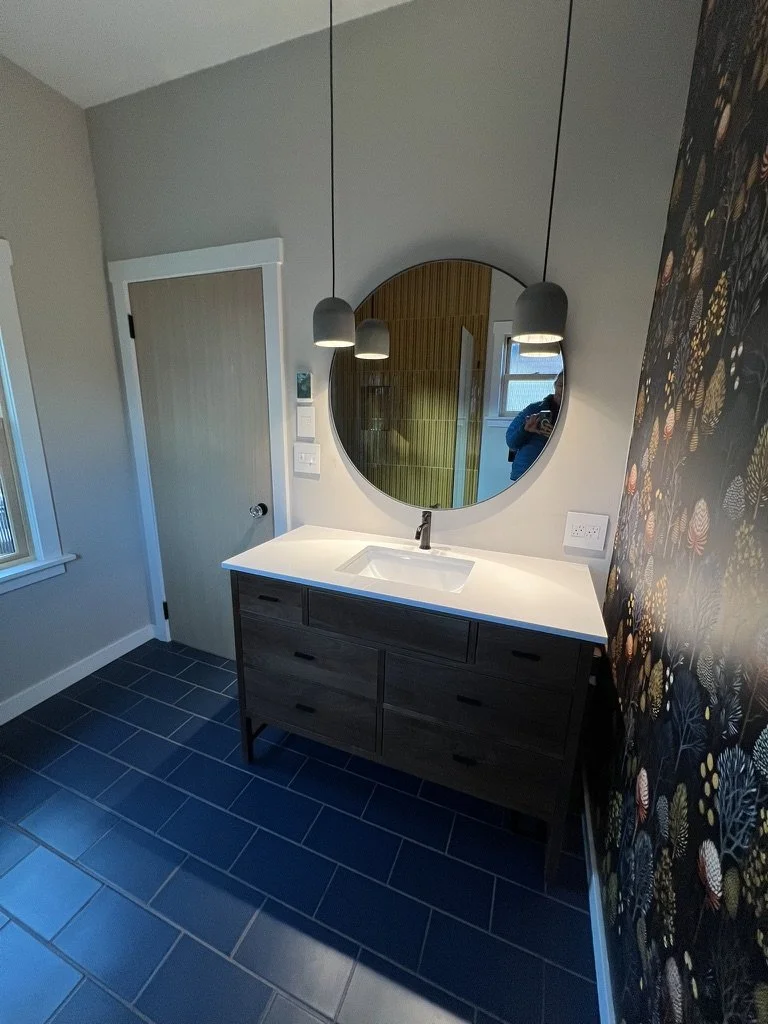 A modern bathroom featuring a dark wooden vanity with six drawers, a white countertop with a built-in sink, a round mirror above the sink, three hanging pendant lights, a light-colored door, and a window beside the vanity. The floor has dark tiles, a