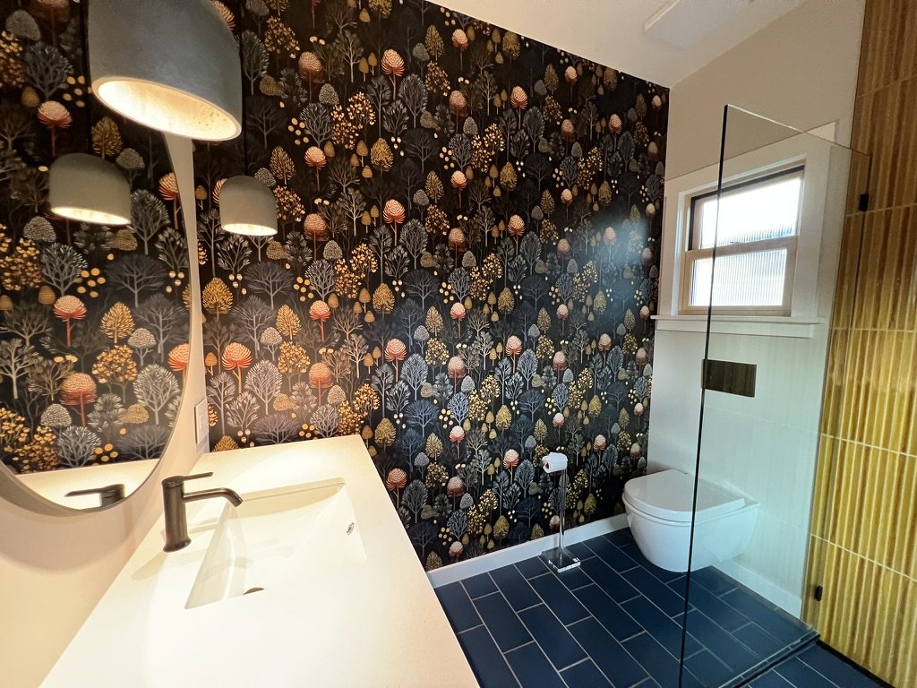 Modern bathroom with a floral patterned wallpaper, a white sink with a faucet, a round mirror, a toilet, and a glass shower enclosure with a window.